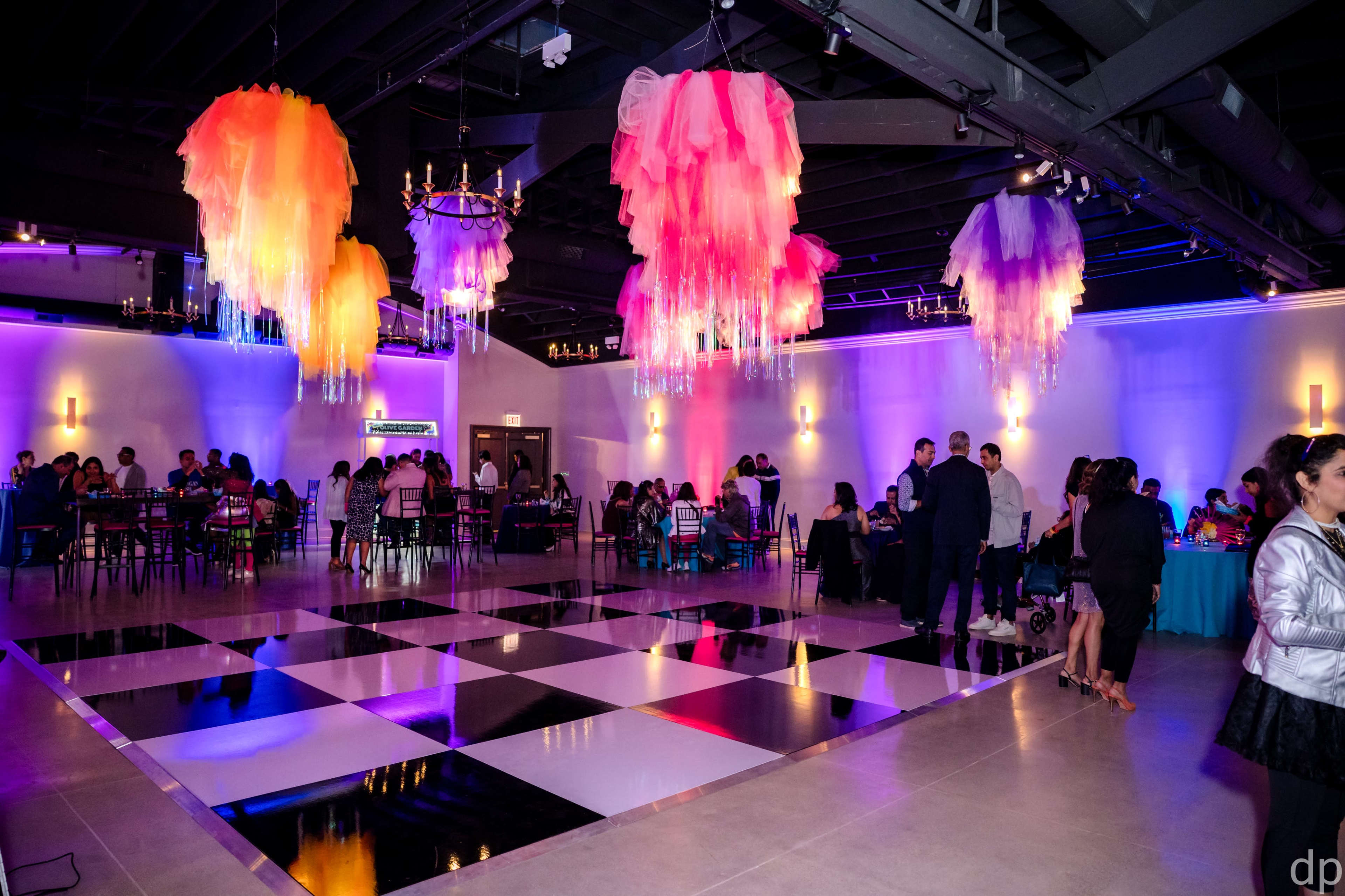The image shows a large event space with a checkered floor, colorful hanging decorations, and guests mingling at tables.