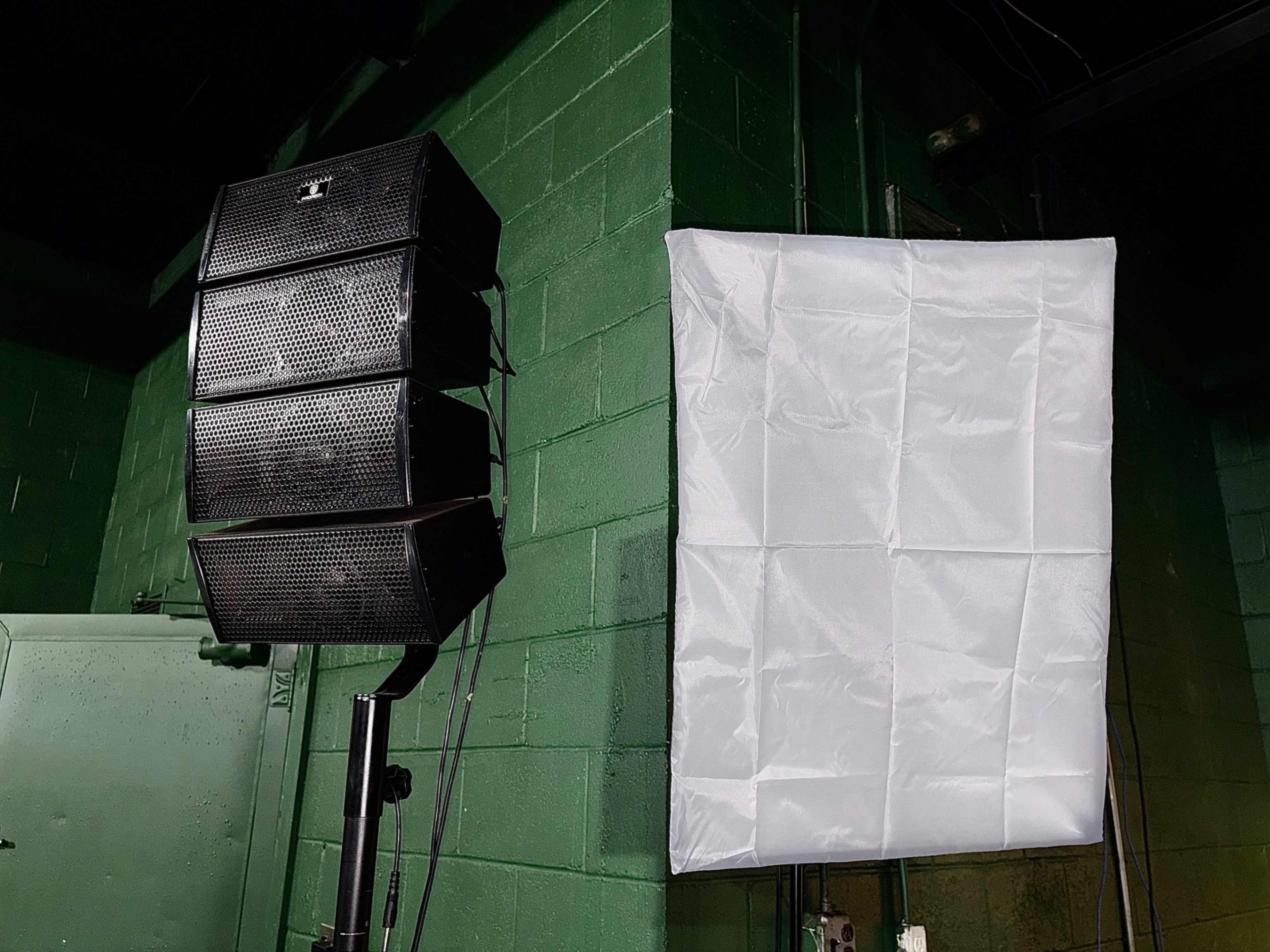 A stack of black speakers is mounted on a pole beside a white diffusion fabric attached to a green wall.