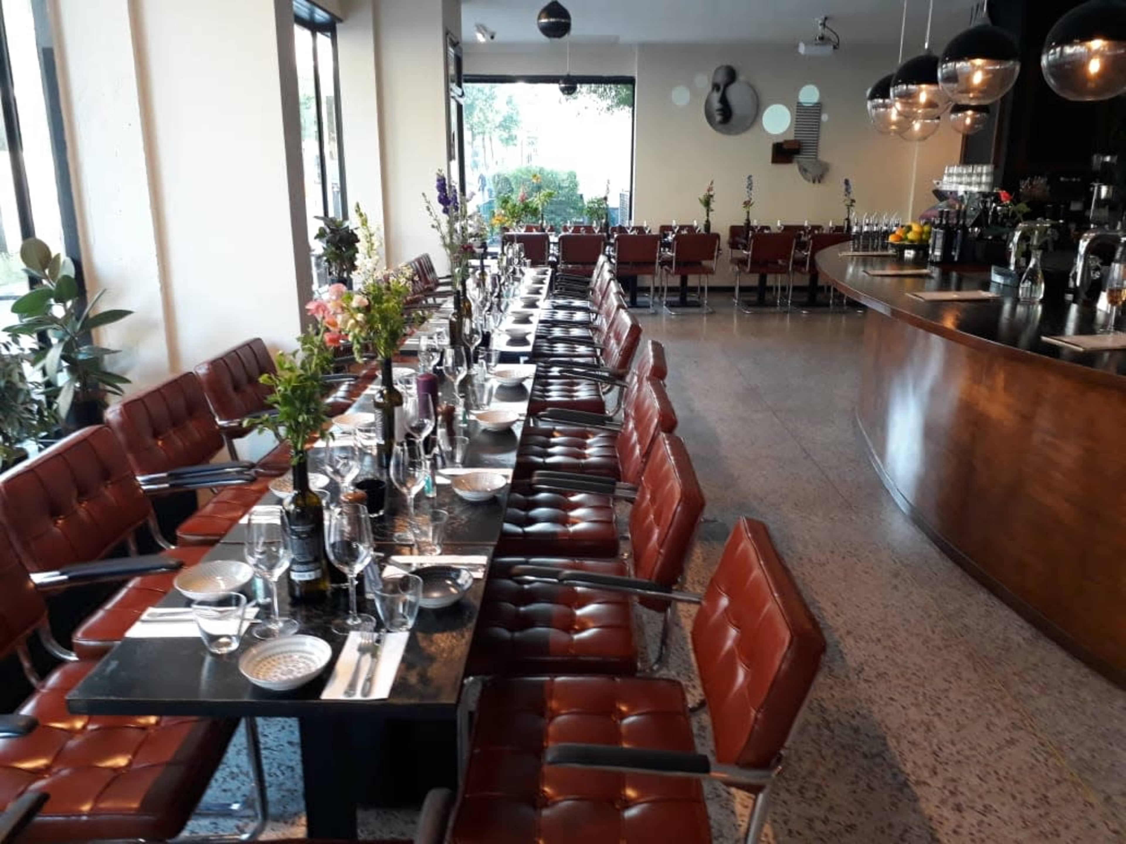 A long dining table with red upholstered chairs is set for a meal in a modern restaurant with large windows and a wooden bar area.