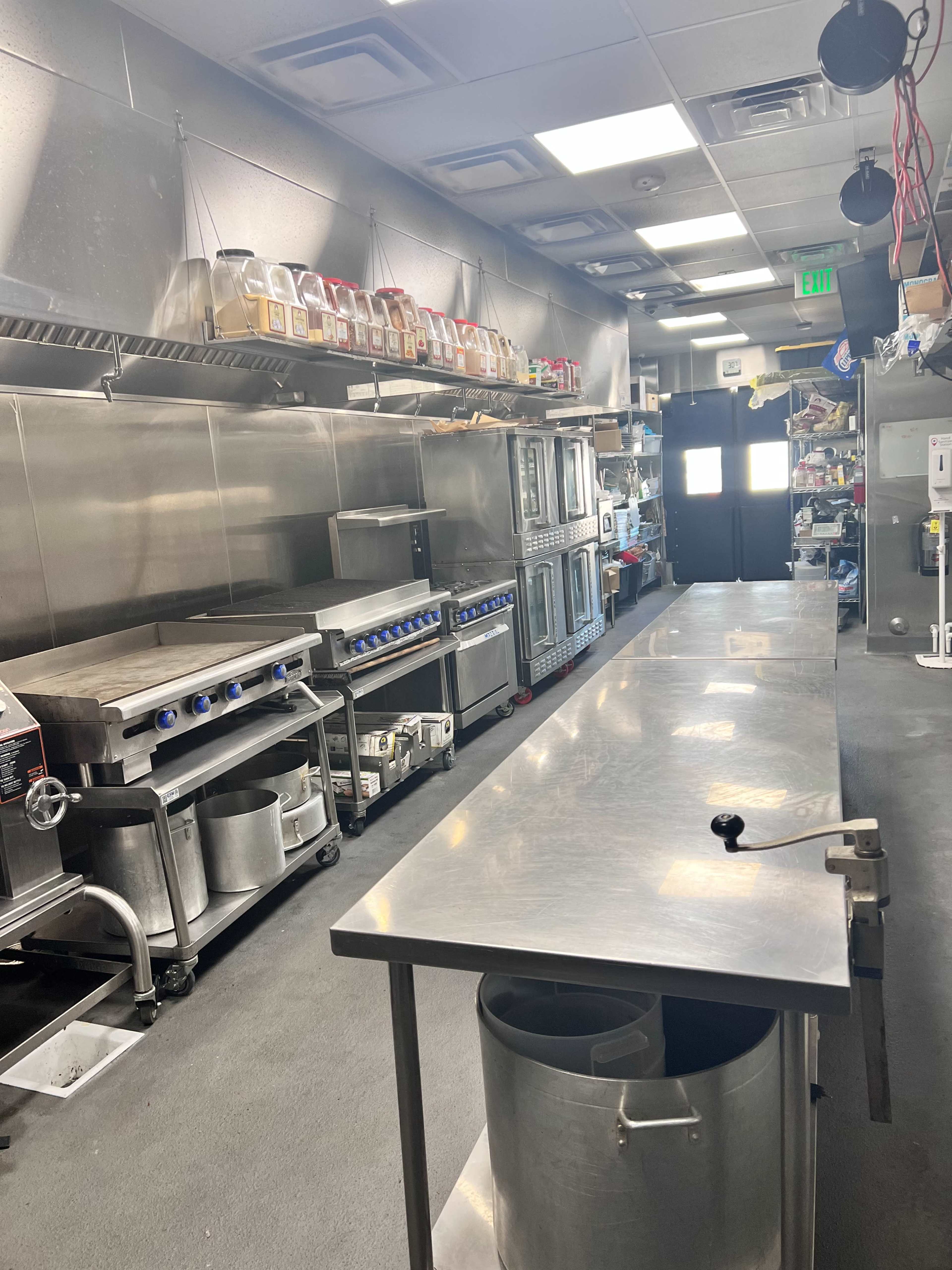 The image shows a commercial kitchen equipped with stainless steel appliances, a long prep table, and storage shelves filled with various containers.