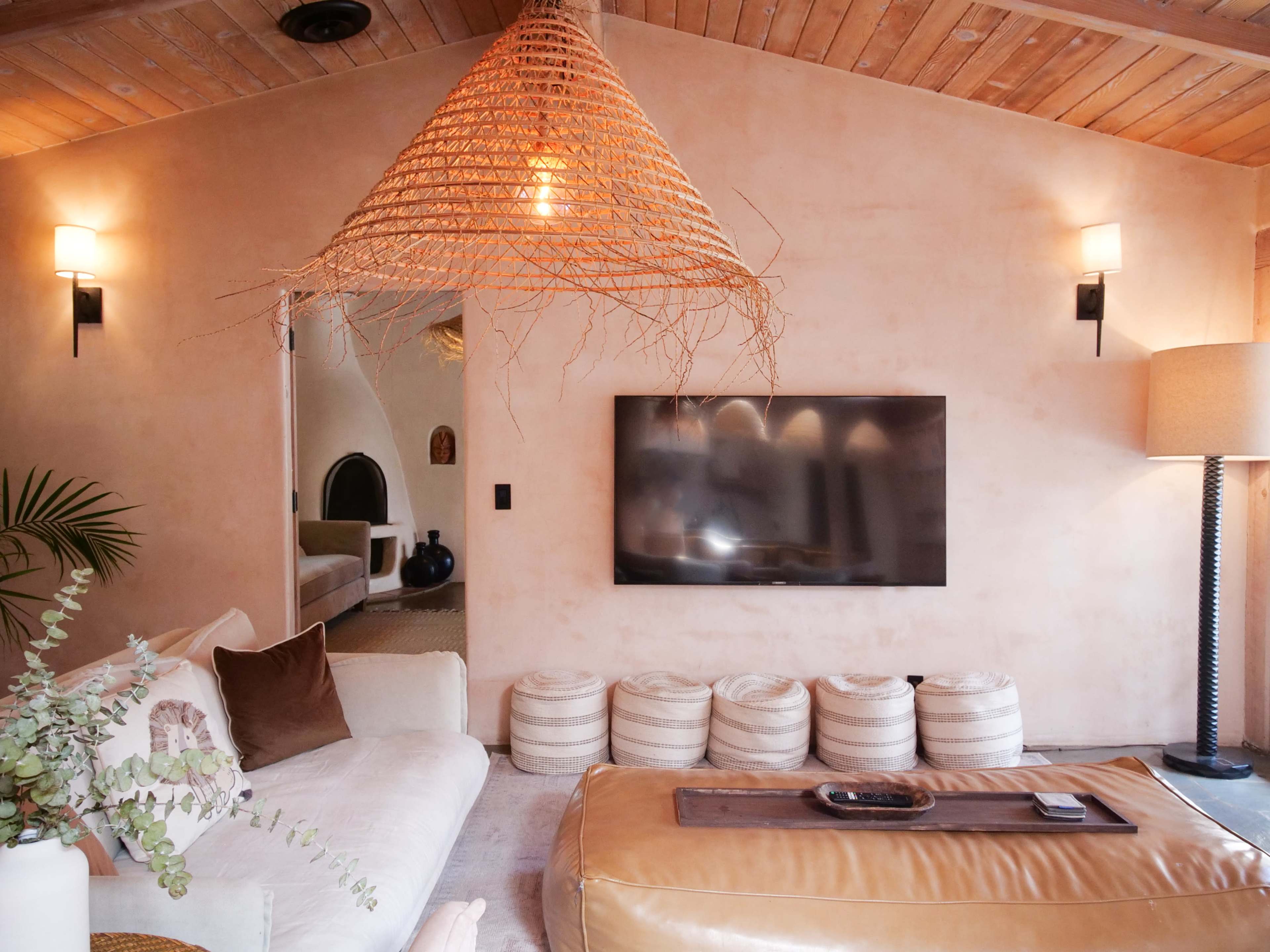 A cozy living room featuring a minimalist style with a large TV mounted on the wall, a light-colored sofa, a textured pendant light, and circular poufs arranged on a rug.