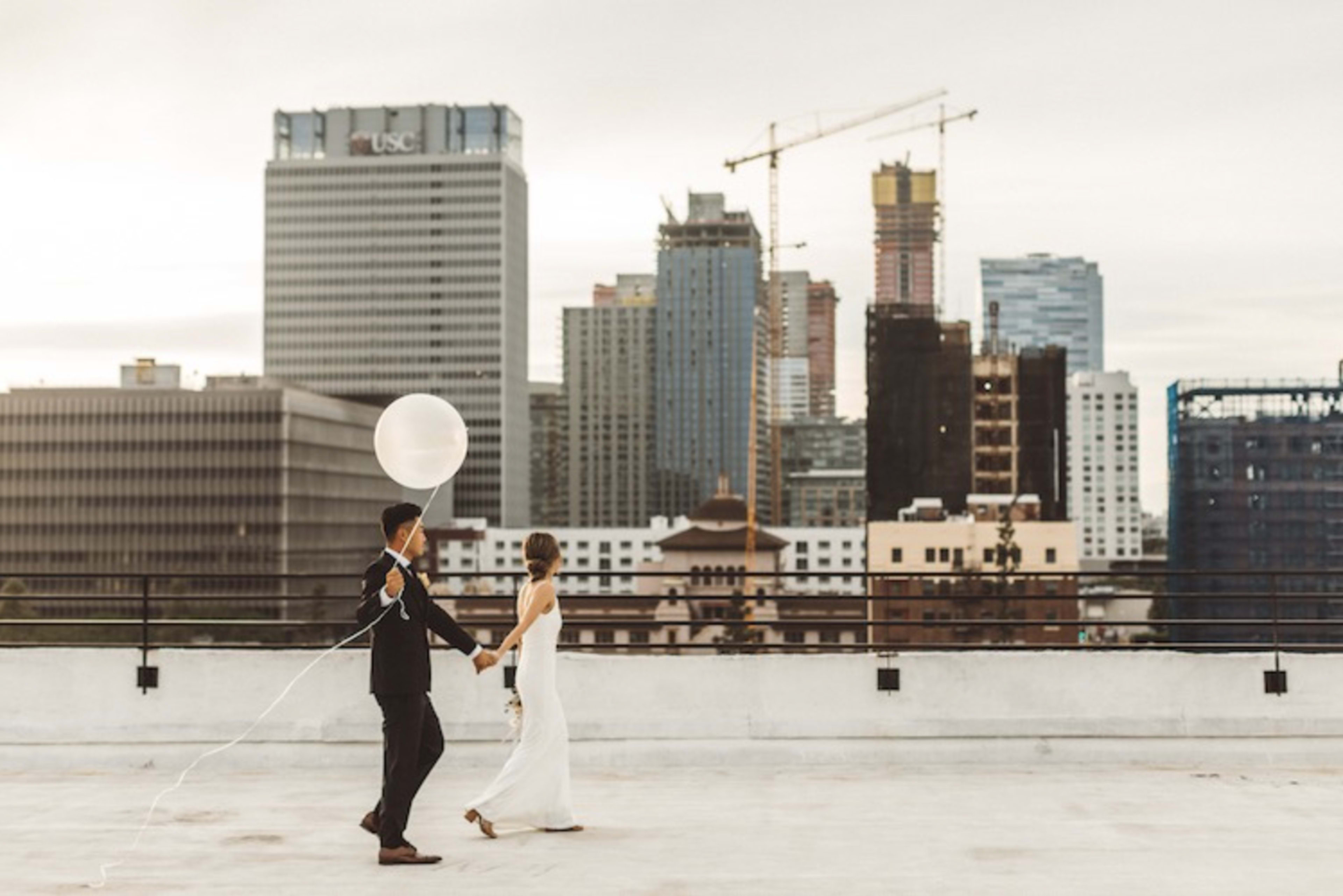 A couple in formal attire walks hand in hand on a rooftop with a city skyline in the background, holding a large white balloon.