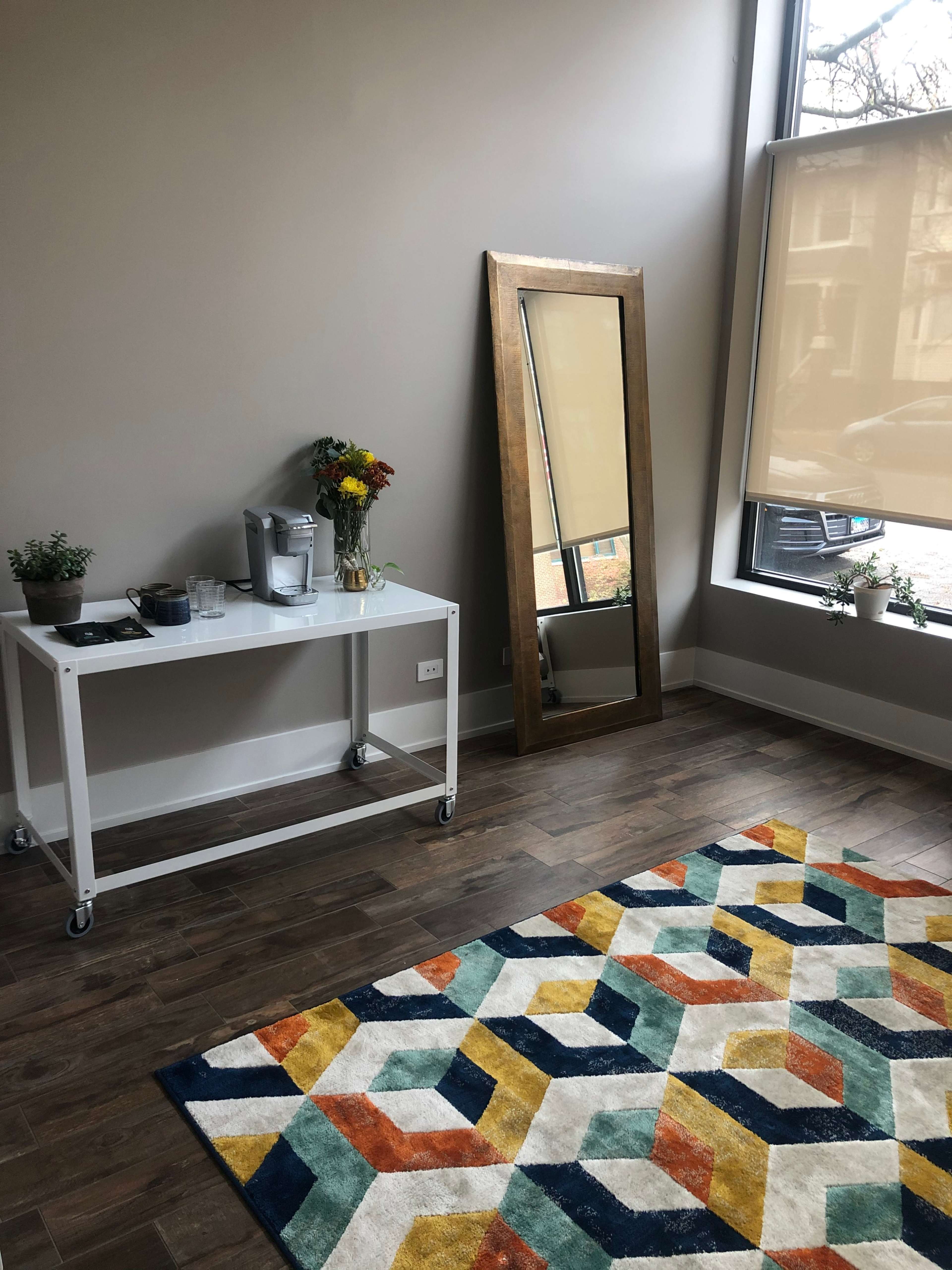 A room with a decorative rug, a mirror leaning against the wall, a small table with a coffee maker, and potted plants.
