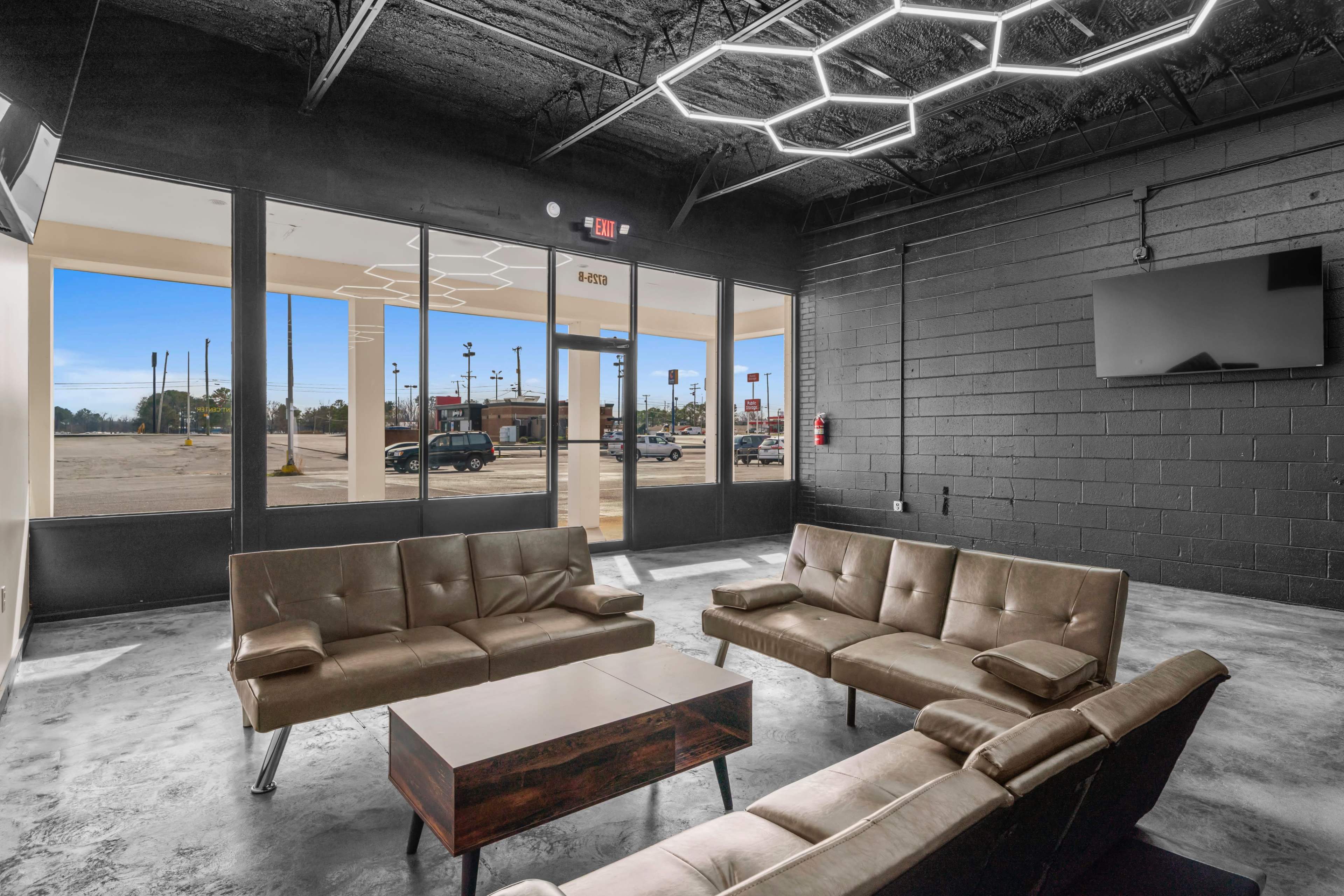 The image shows a modern lounge area with brown leather couches and a coffee table, facing large windows that overlook a parking lot and street.