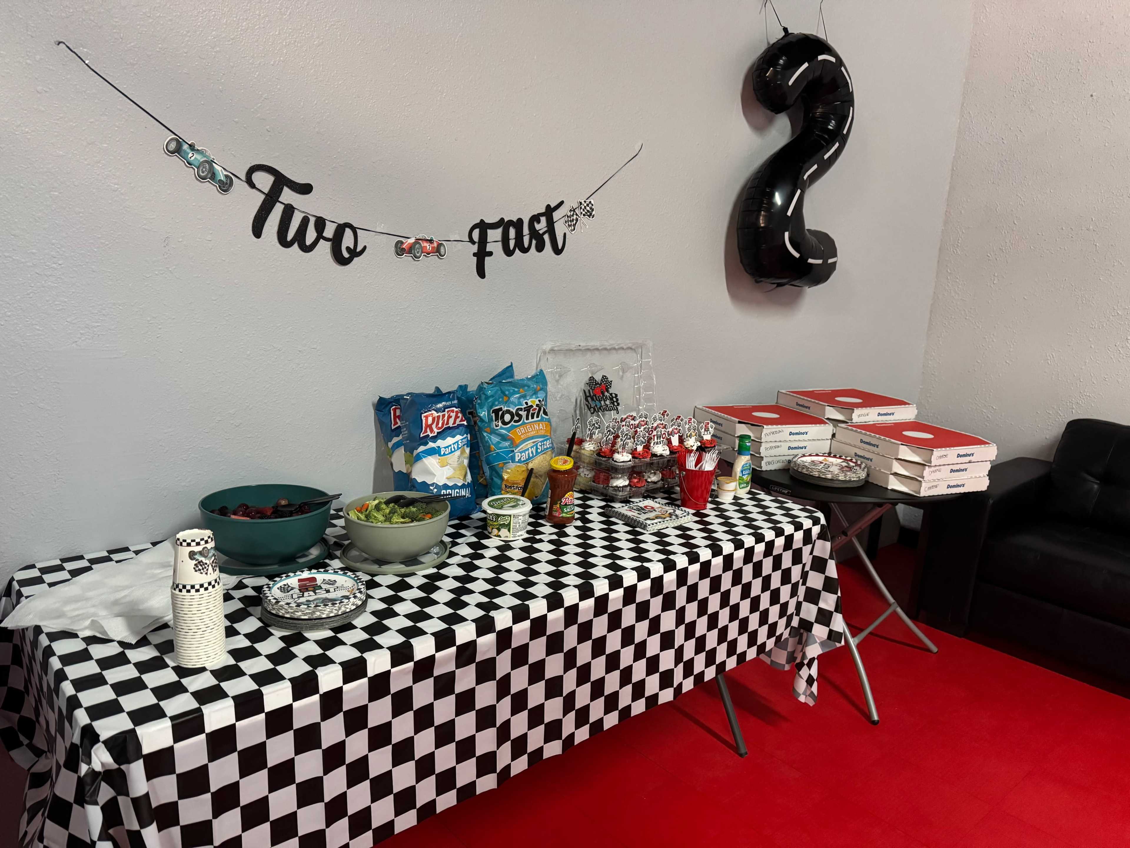 The image shows a party setup with a checkered tablecloth displaying snacks, salads, and pizzas, accompanied by decorations that celebrate a second birthday.