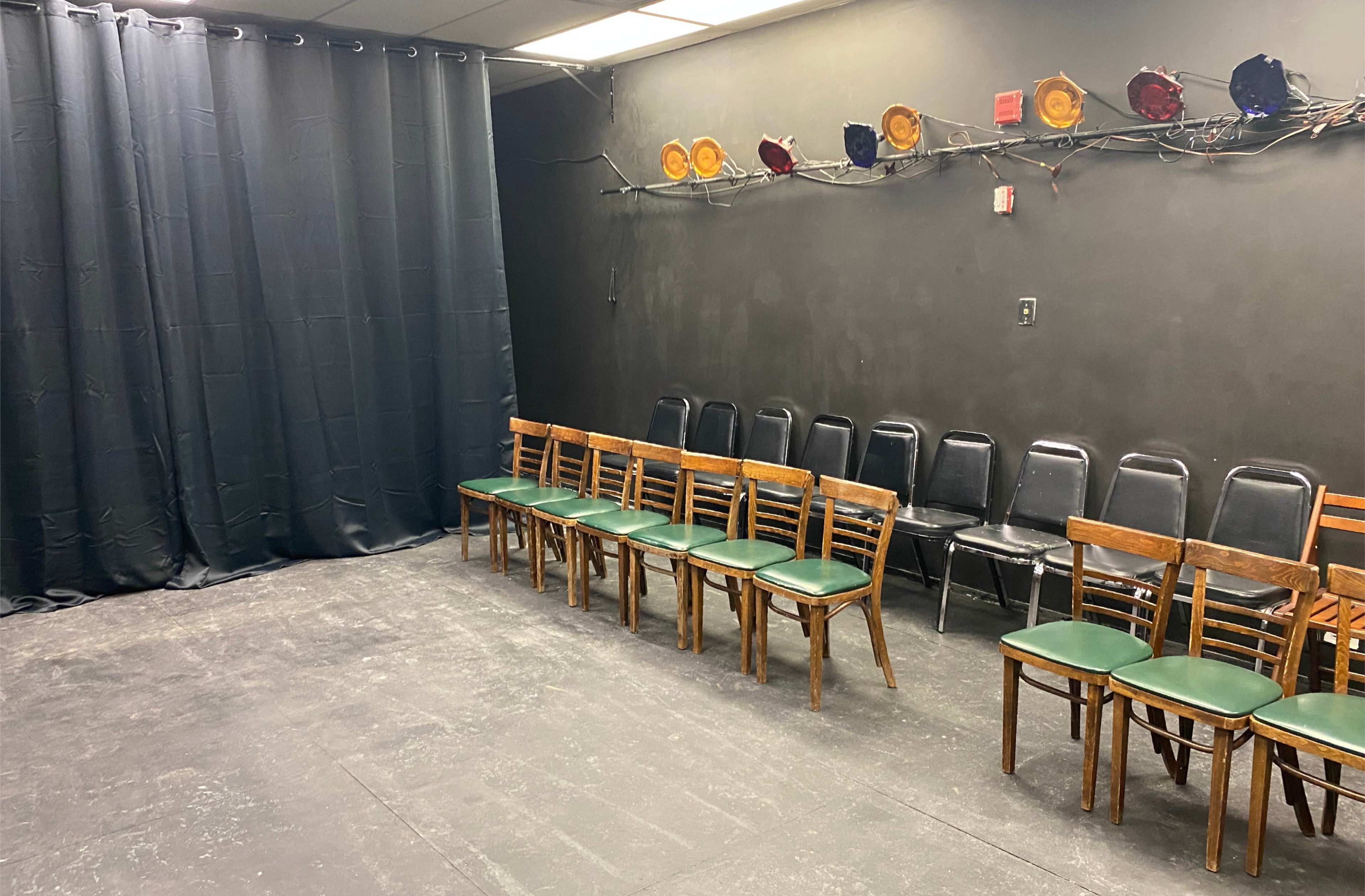 The image shows a dimly lit rehearsal space with a row of wooden chairs on one side and a black curtain on the other side, illuminated by overhead stage lights.
