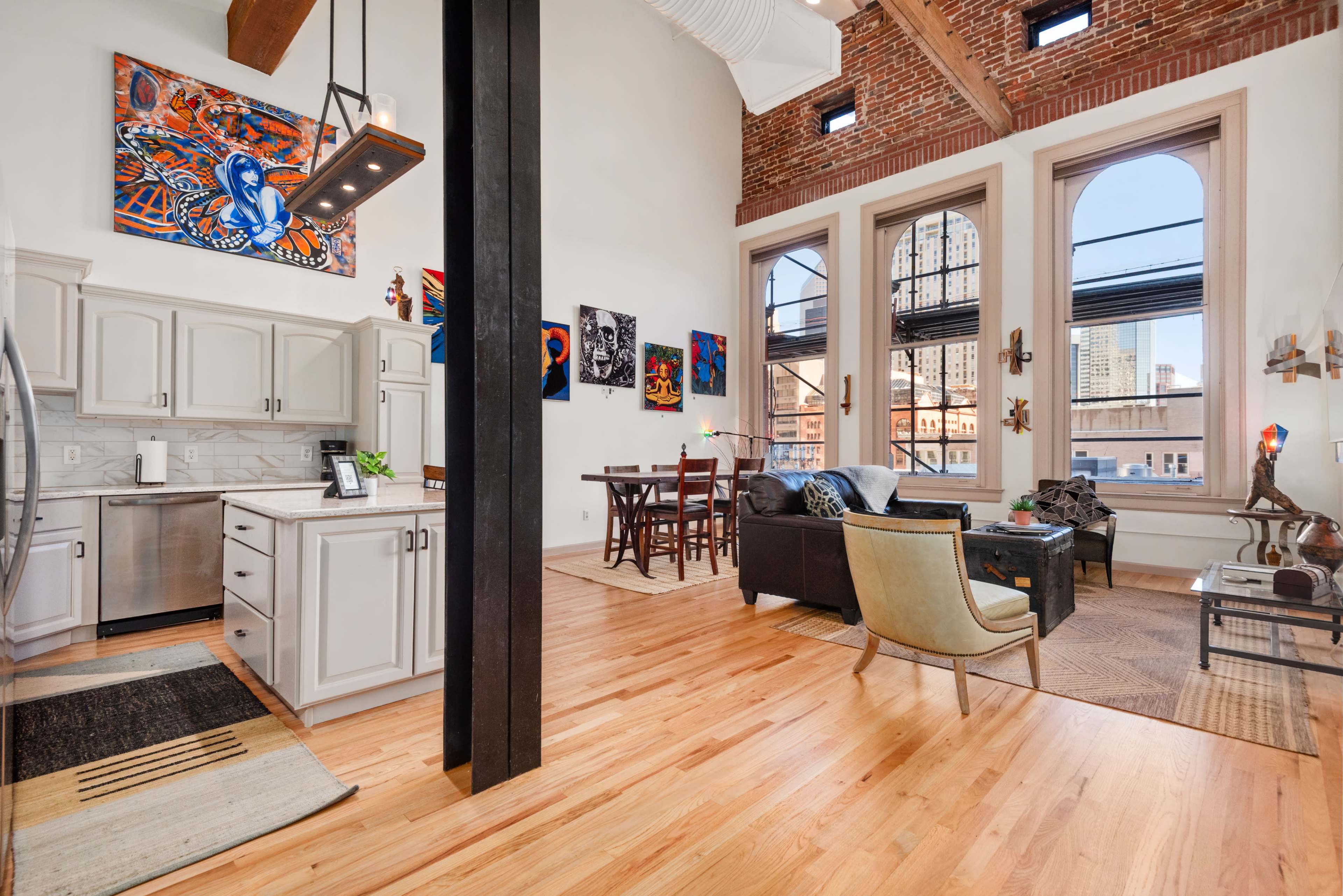 The image shows a spacious, open-concept living area featuring a modern kitchen with stainless steel appliances, a dining area with wooden furniture, and a living room with a couch and decorative art on the walls.