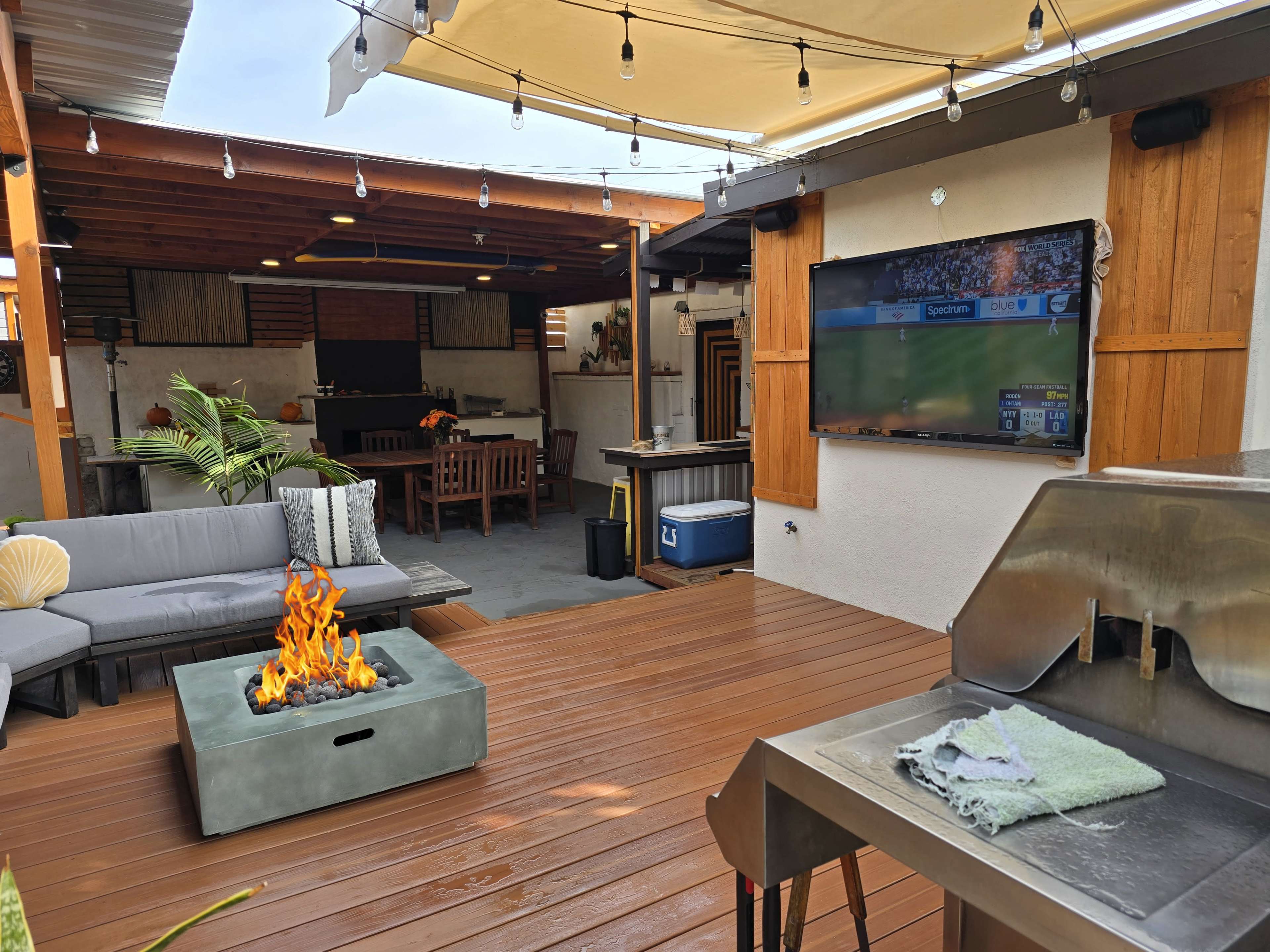 The image shows a backyard patio featuring a seating area with a fire pit, a grill, and a television mounted on the wall.