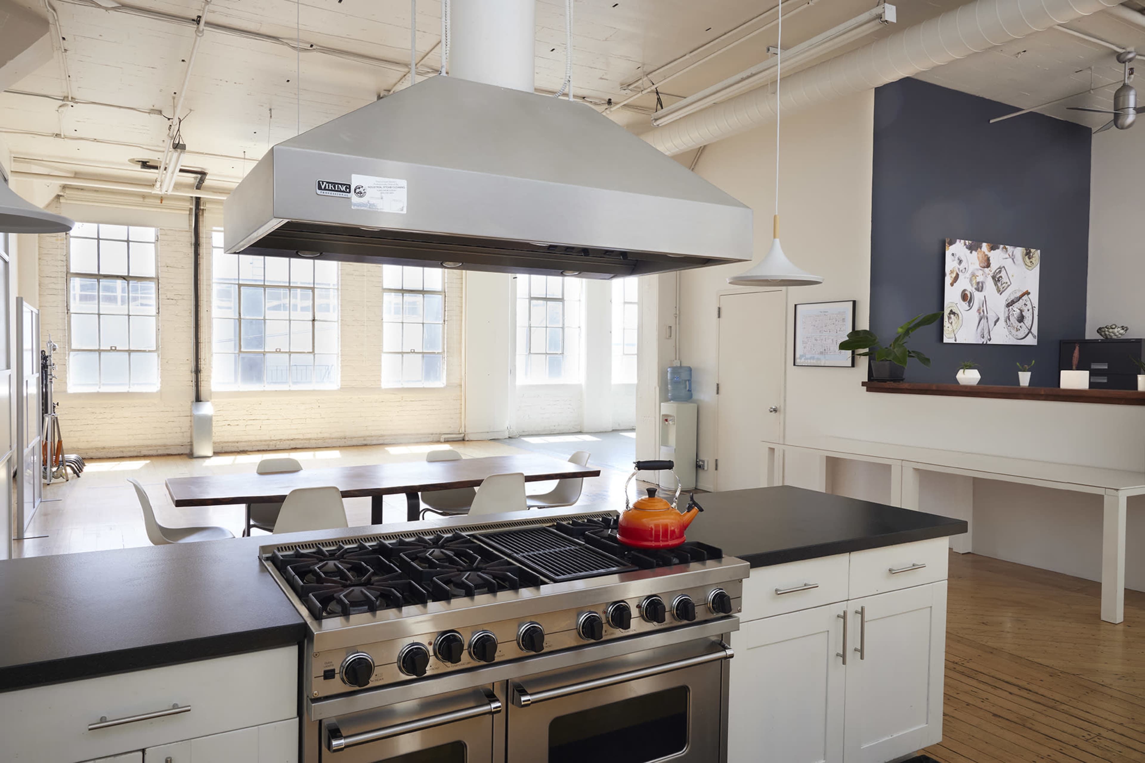 A modern kitchen with a stainless steel hood, a gas stove, and a dining area featuring a long table and chairs, illuminated by natural light from large windows.