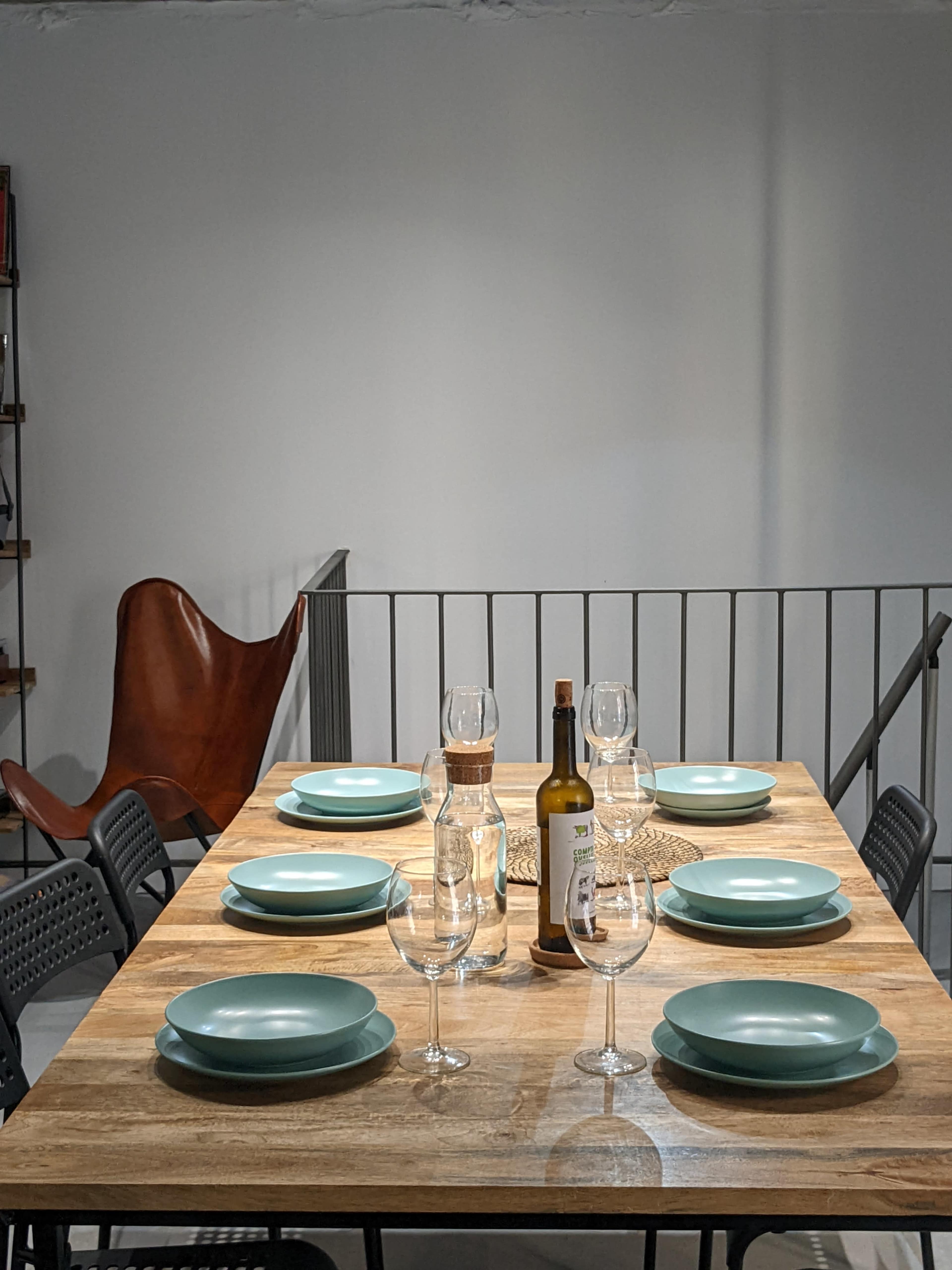 A dining table is set with six turquoise plates and wine glasses, accompanied by a bottle of wine and water, in a minimalist room featuring an accent chair.