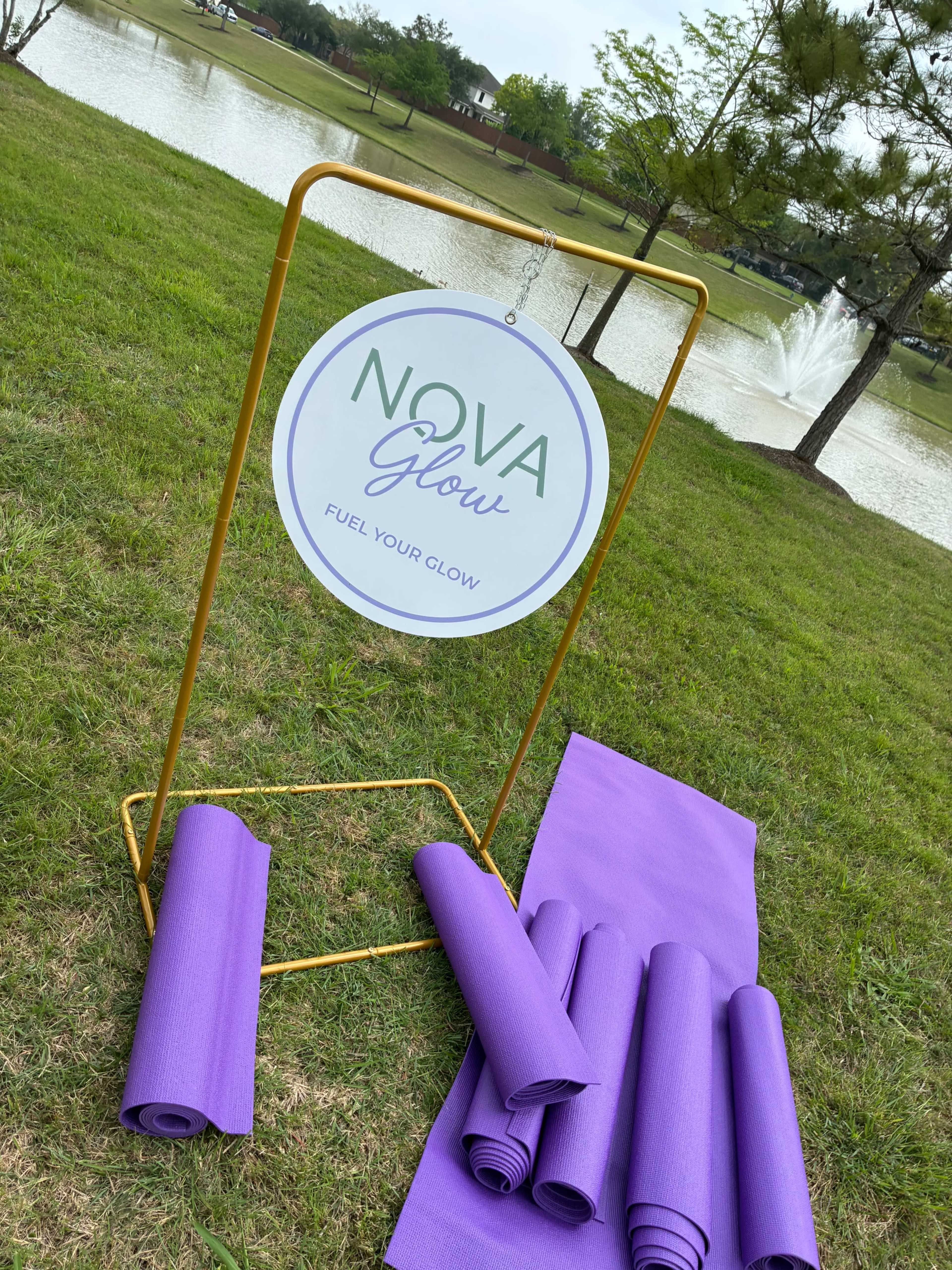 A sign for "NOVA Glow" stands next to neatly rolled purple yoga mats on grass by a pond with a fountain in the background.