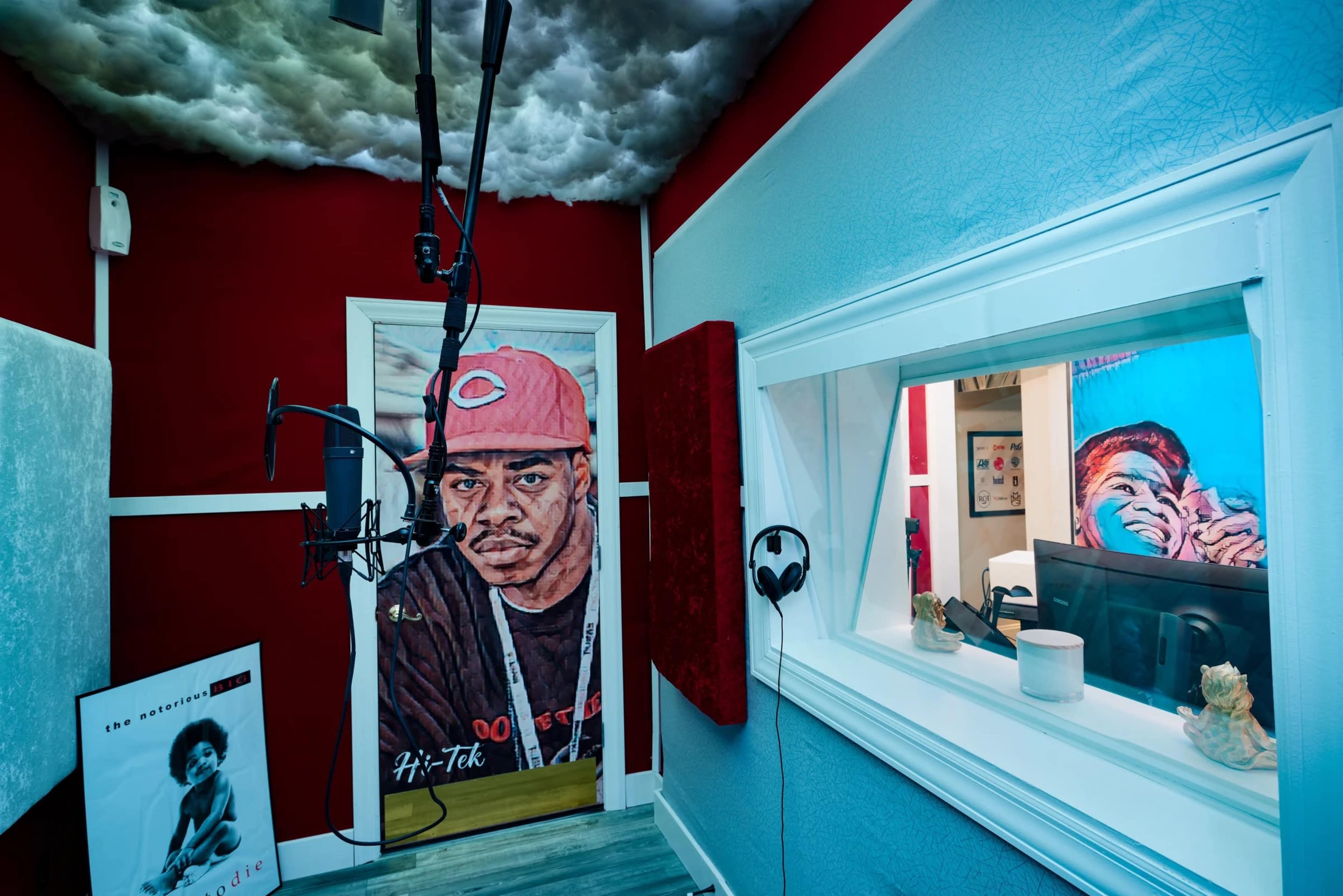 The image shows a recording studio booth featuring a microphone, headphones, and a mural of a man wearing a red cap on the door.