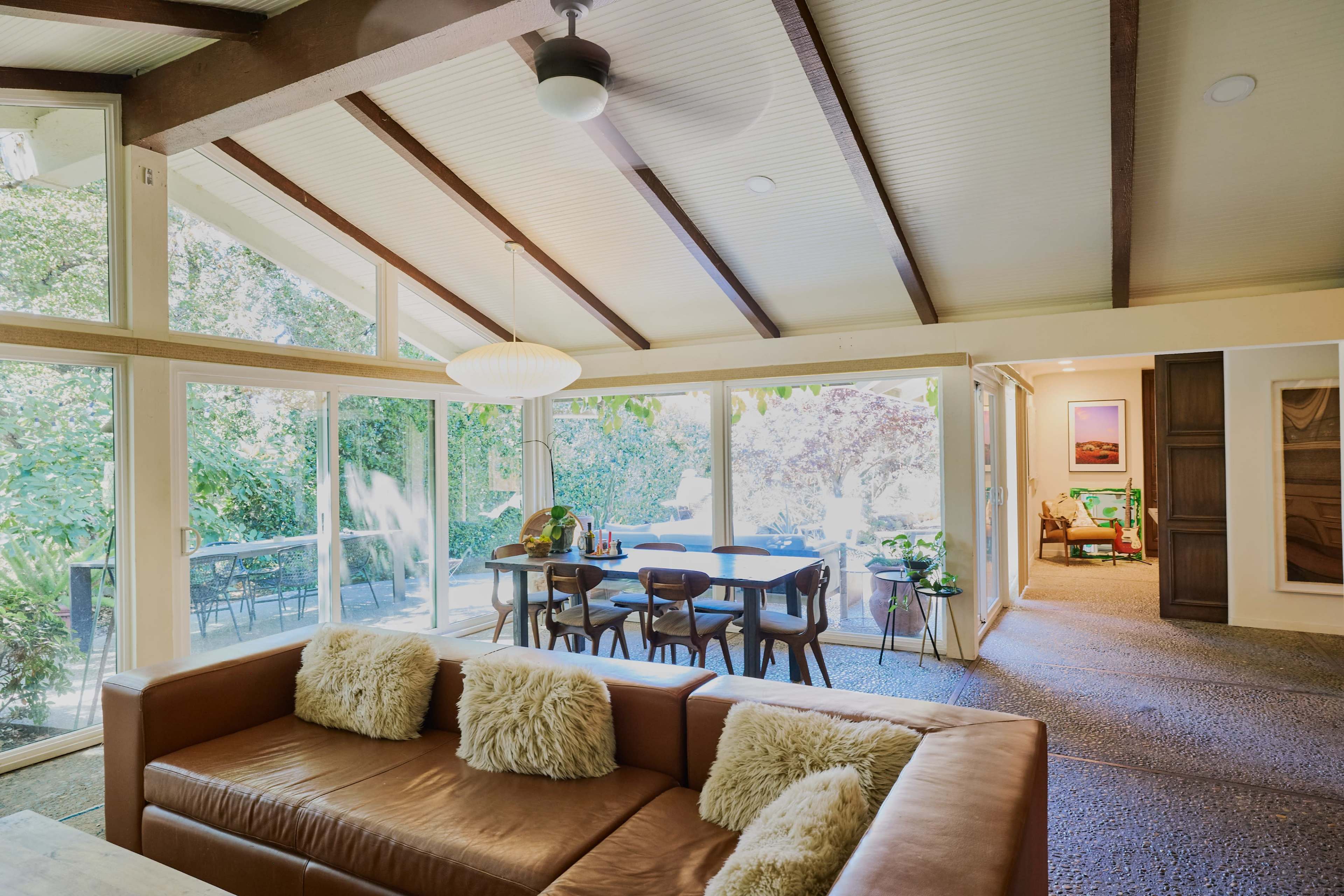 A living room with a brown leather sofa and a dining area, featuring large glass doors that open to an outdoor space with greenery.