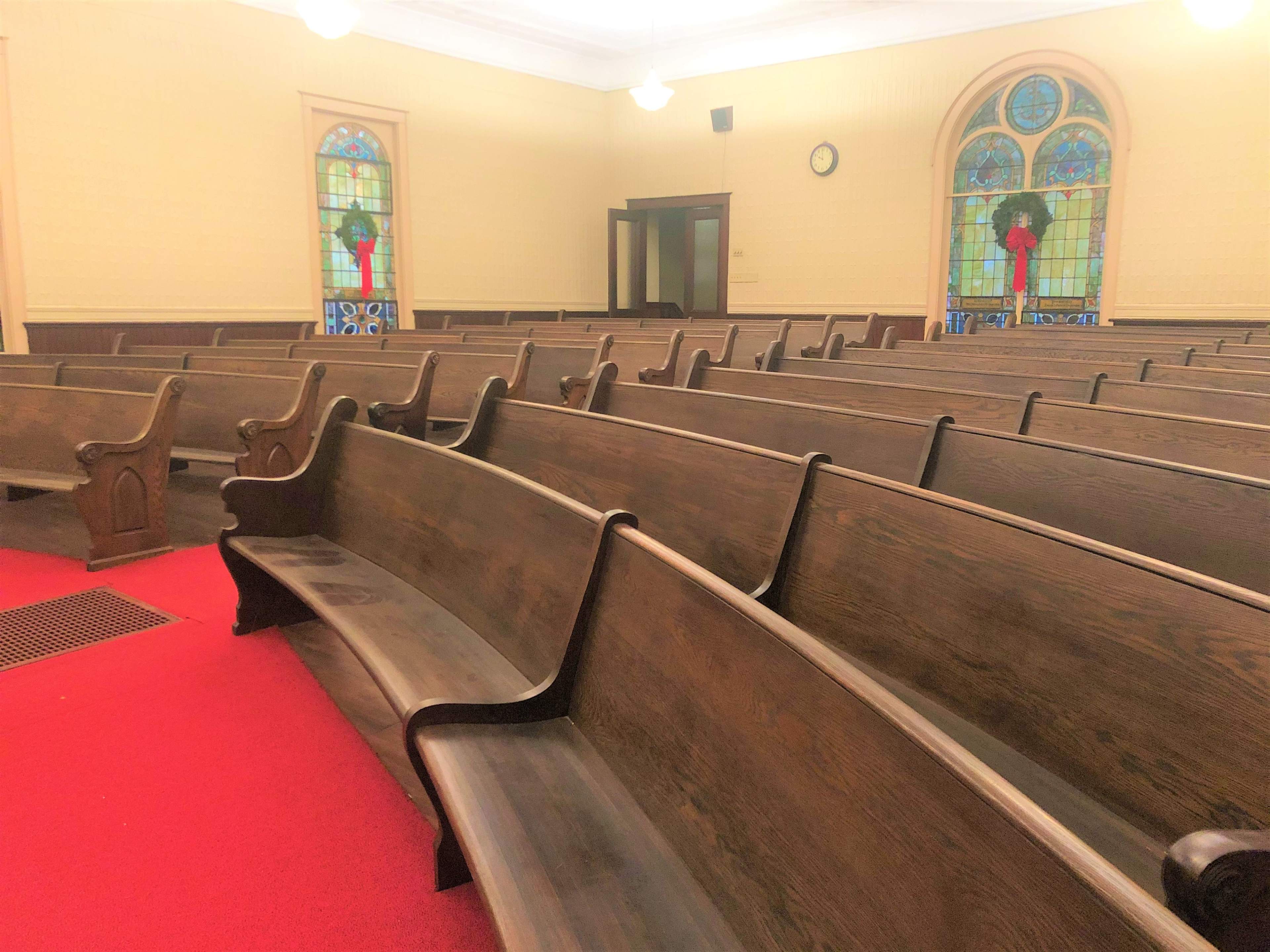 A row of wooden pews arranged on a red carpet inside a church, with stained glass windows in the background.