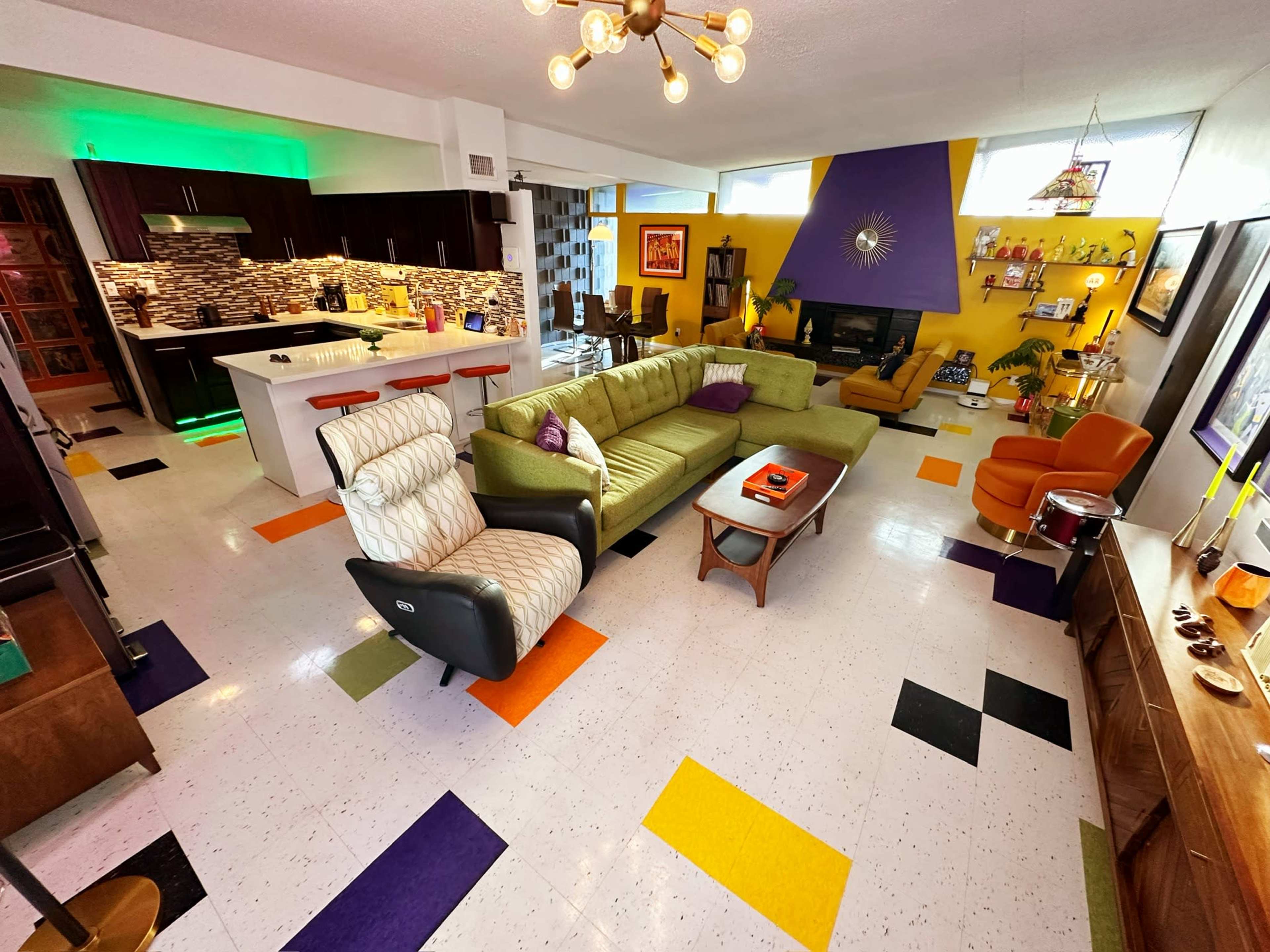 The image shows a colorful retro-inspired living room with a sectional sofa, armchair, and a modern kitchen area in the background.