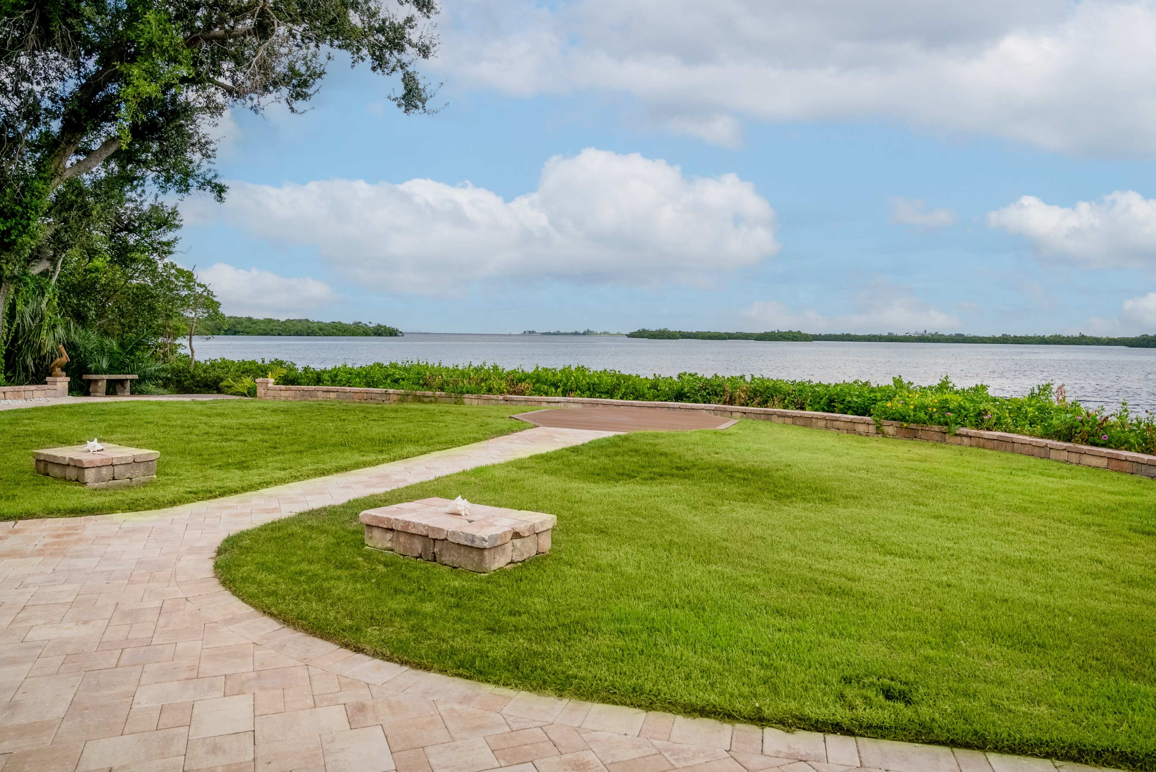 A landscaped area features two stone benches along a curved pathway, overlooking a calm body of water under a cloudy sky.