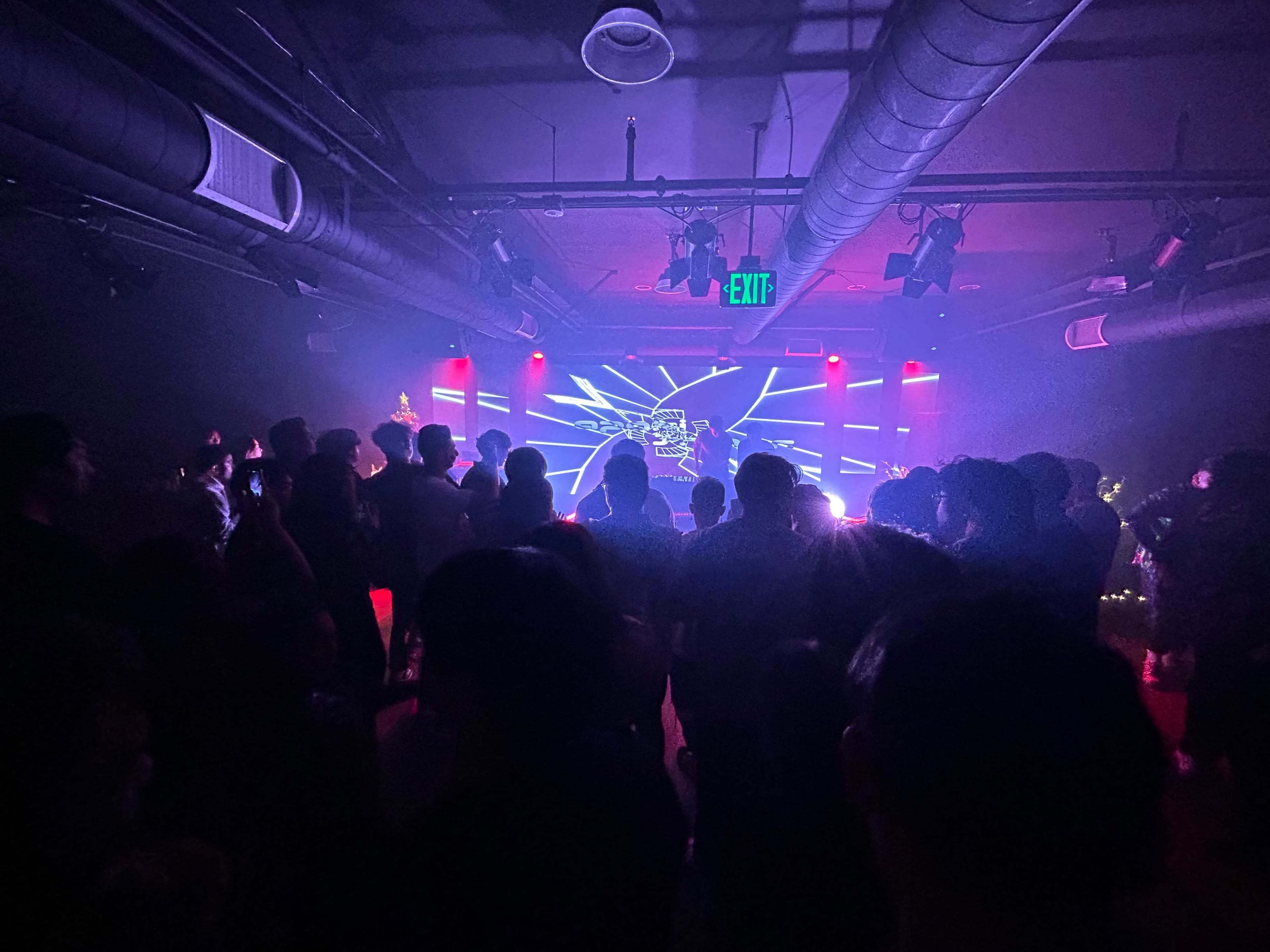 A crowd gathers in a dimly lit venue with colorful lights and visual displays illuminating the stage.