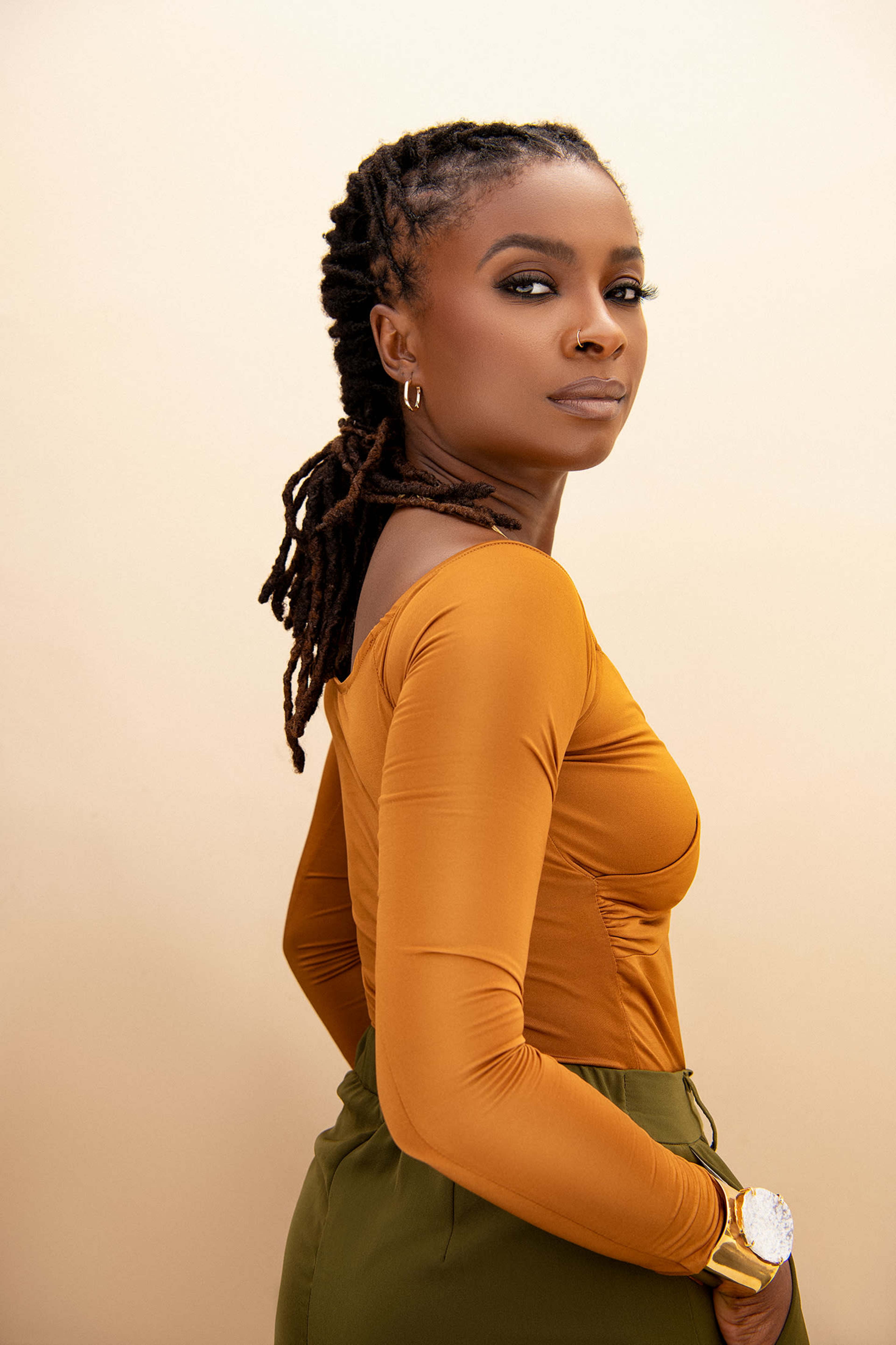 A woman with braided hair is posed against a pale background, wearing a fitted mustard-colored long-sleeve shirt and green pants.