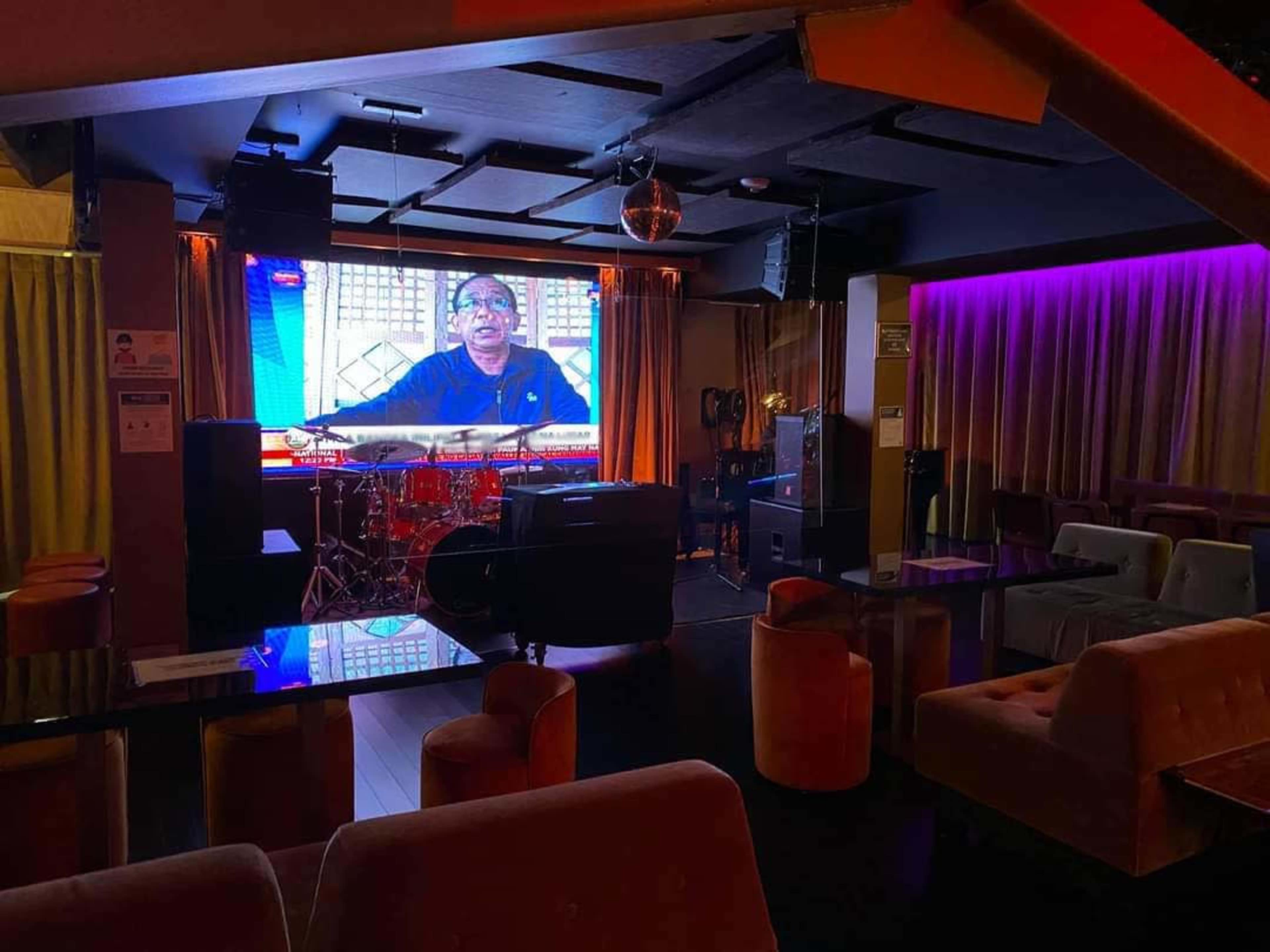 A dimly lit lounge with seating arrangements and a large screen displaying a man, accompanied by a drum set and music equipment.