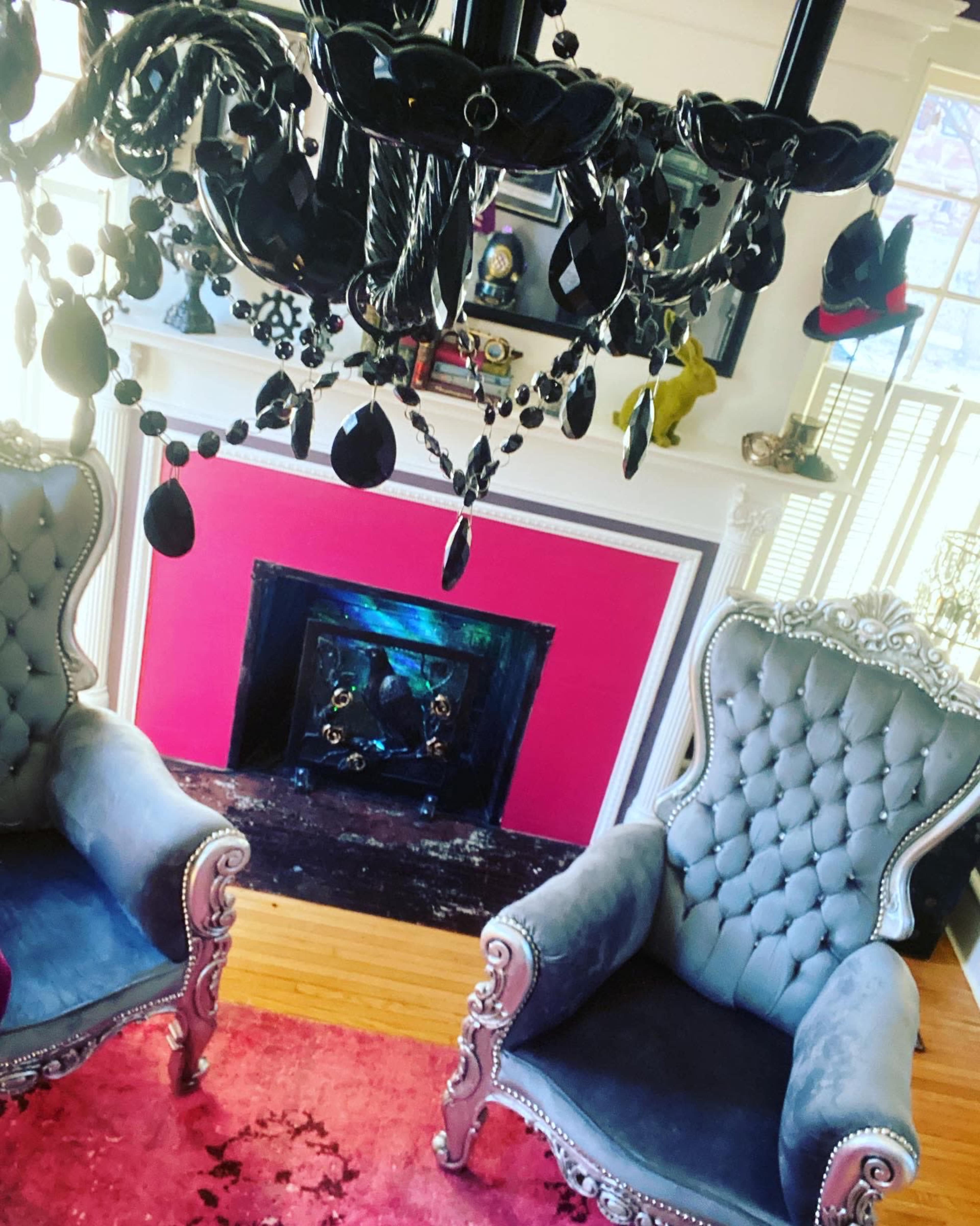 The image shows a living room with two tufted chairs facing a colorful wall, a chandelier above, and a fireplace adorned with decorative items.