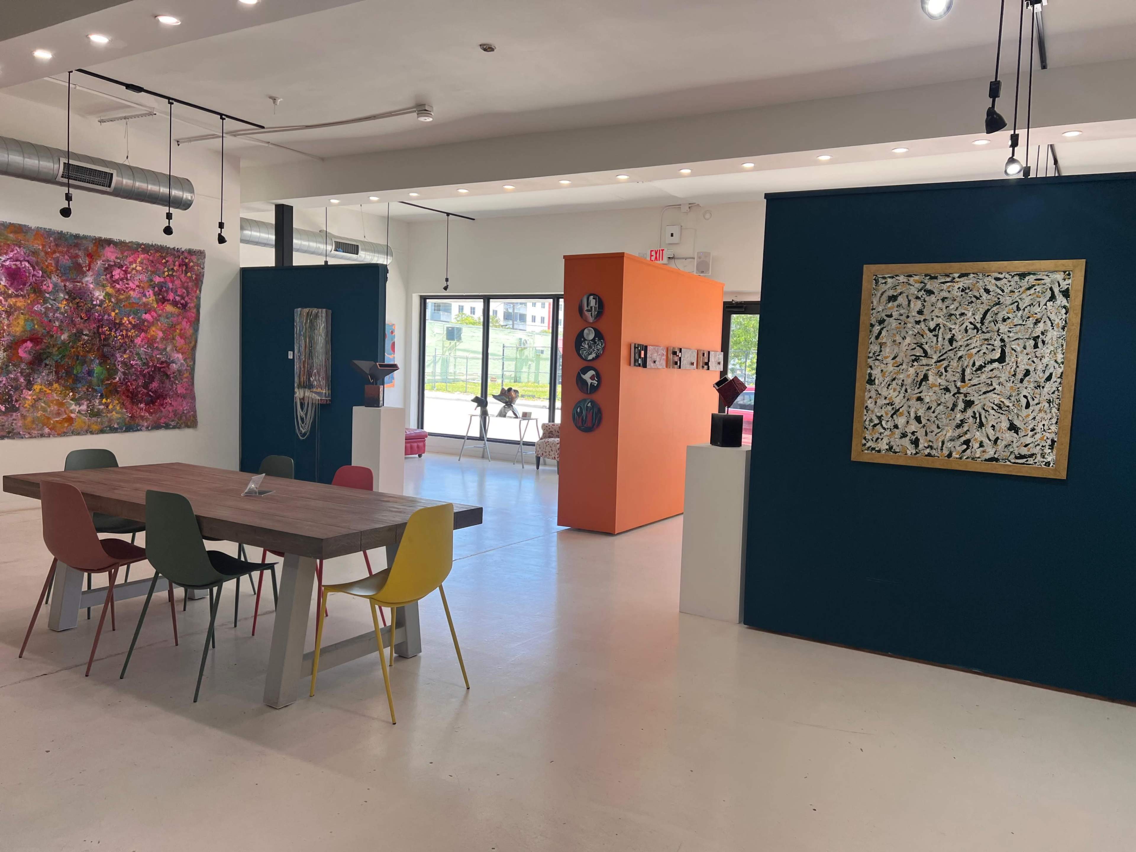 The image shows a modern gallery space featuring a dining table with colorful chairs, vibrant artwork on the walls, and an orange accent wall displaying circular art pieces.