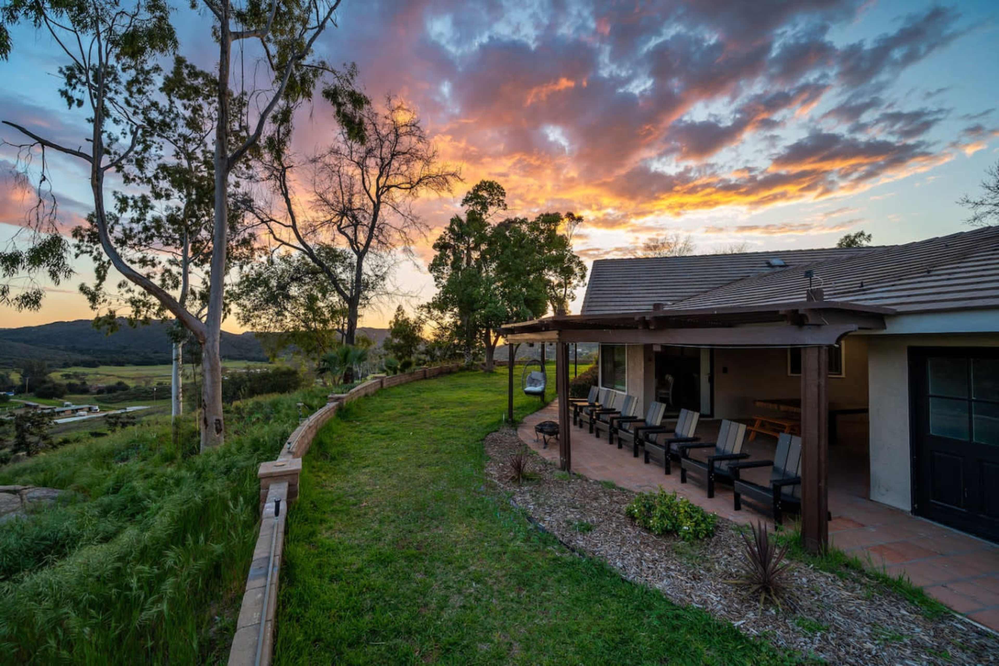 A hillside property features a patio with seating and a scenic view of a sunset over the landscape.