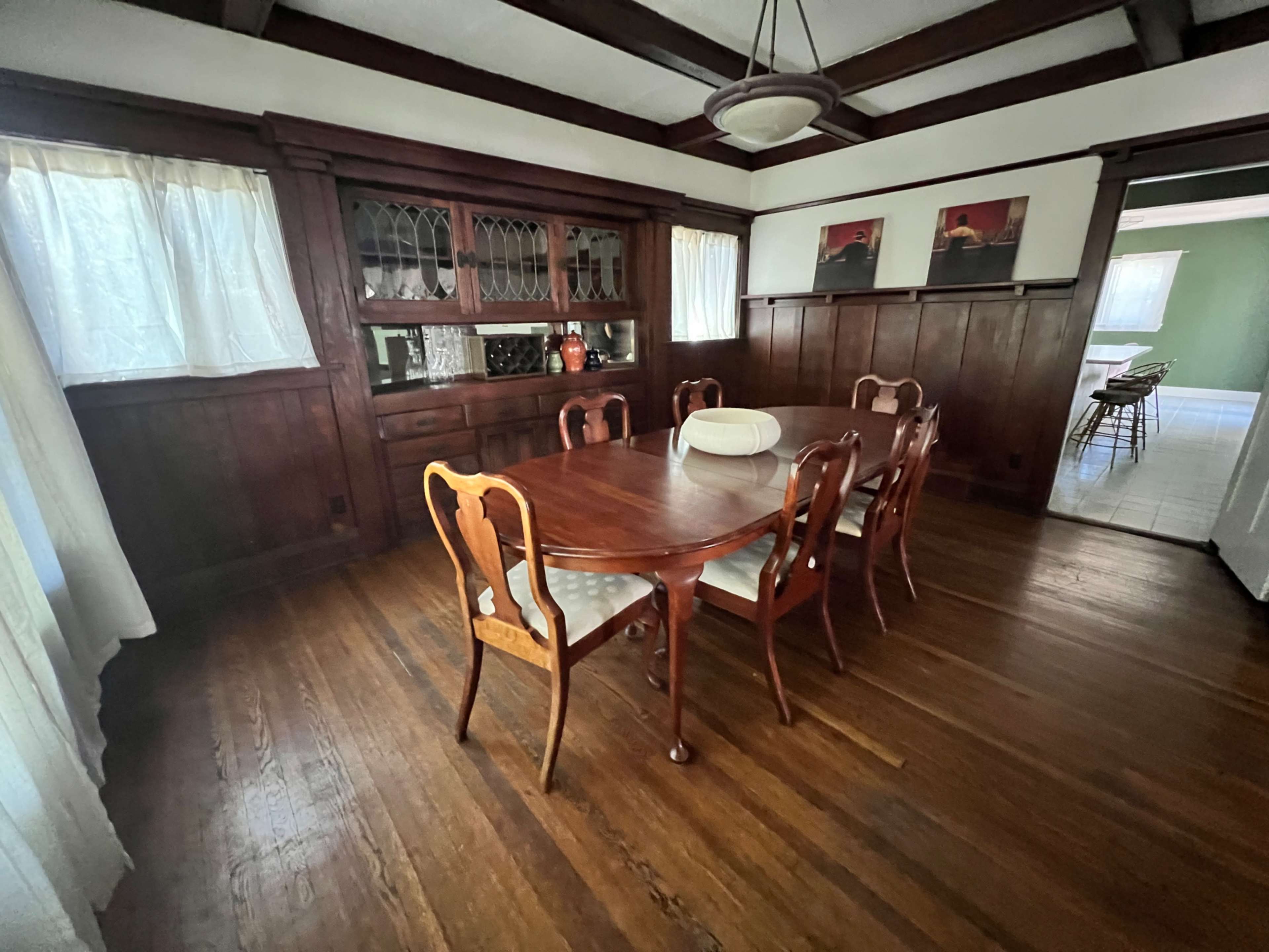 The image shows a dining room with a wooden table and chairs, a china cabinet, and large windows with sheer curtains.