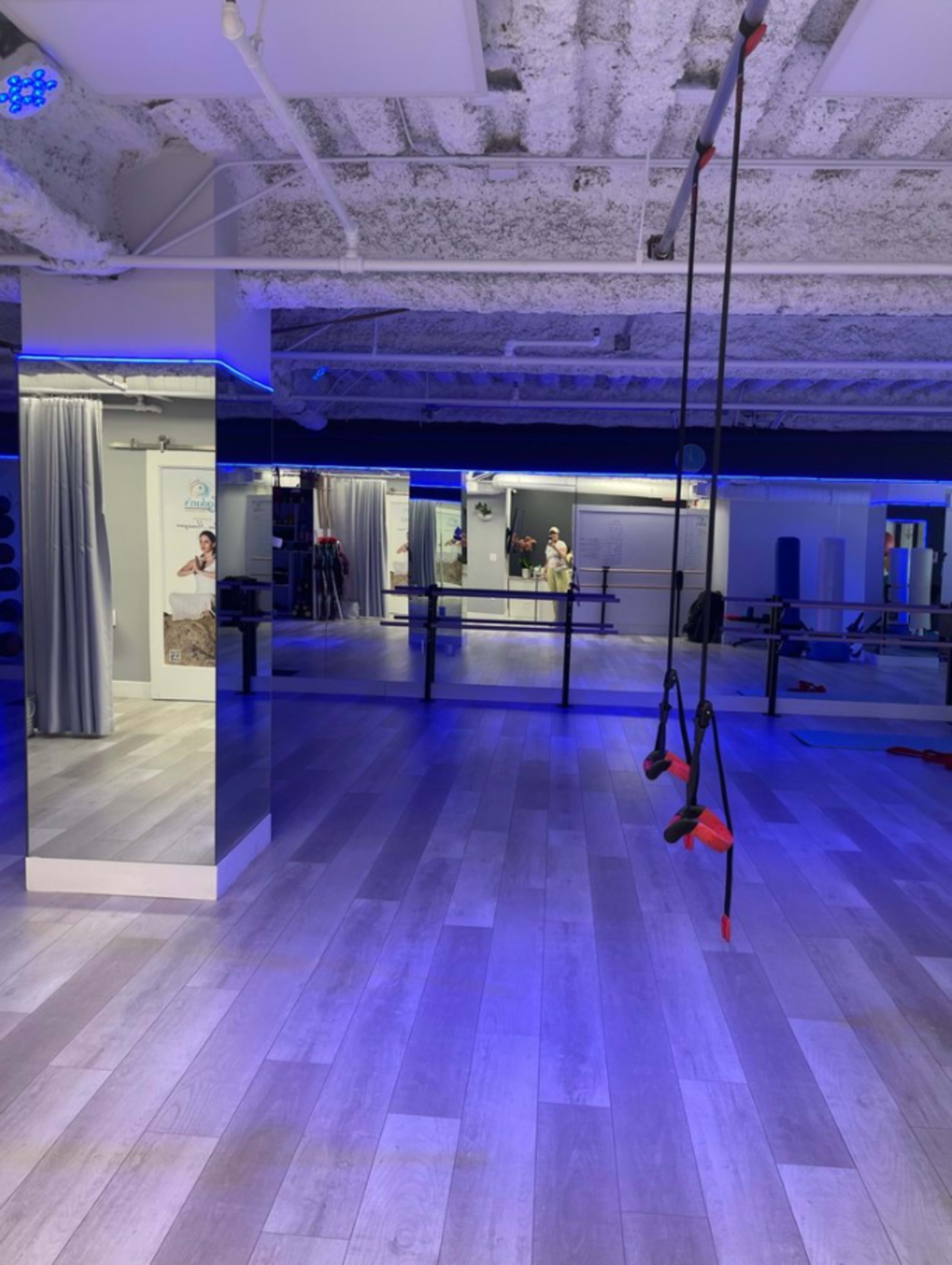 The image shows a fitness studio with mirrors, wooden flooring, and suspension training equipment hanging from the ceiling under blue lighting.