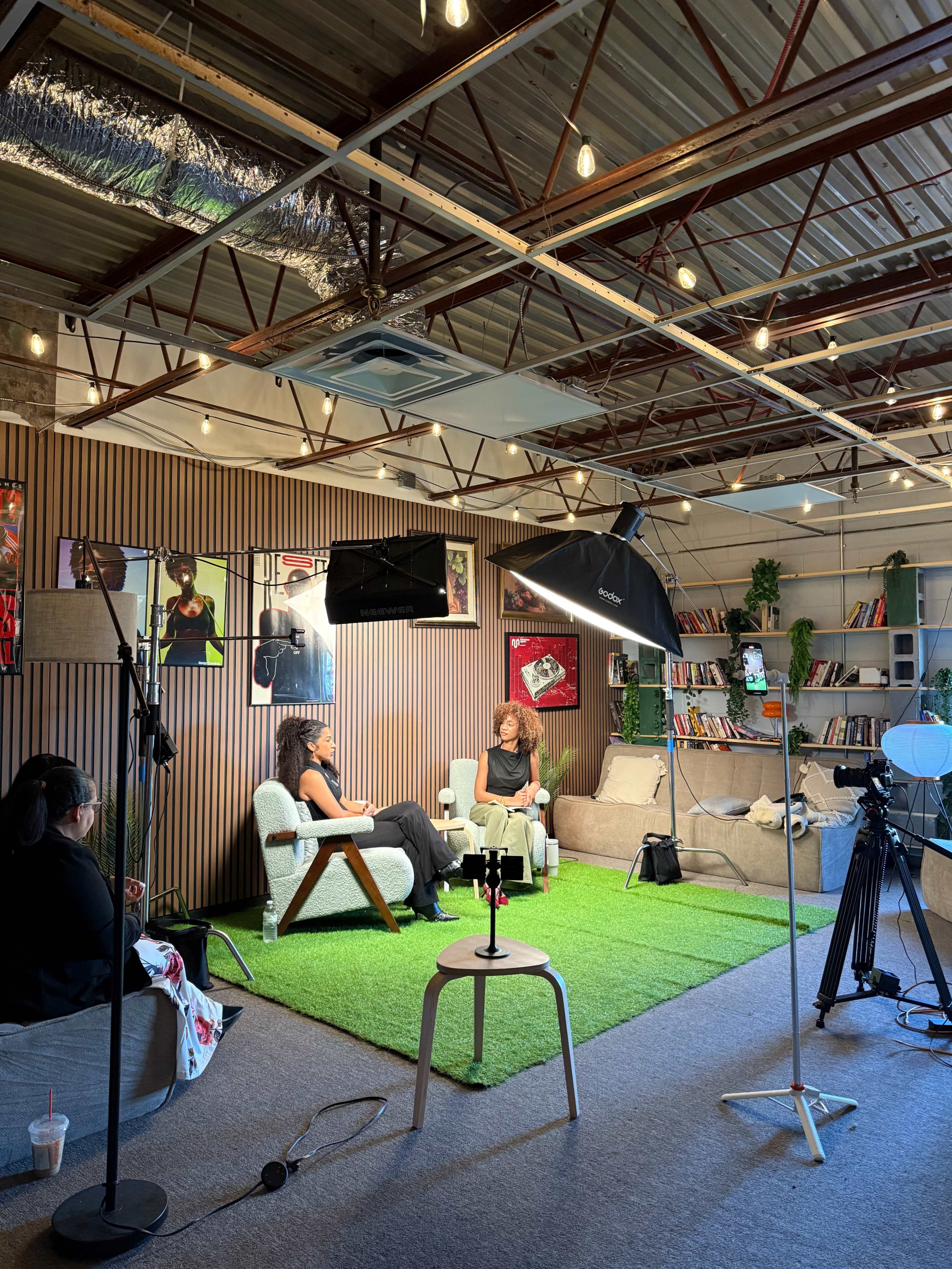 Two individuals are seated on a couch in a modern studio setting, surrounded by bookshelves and professional lighting equipment.