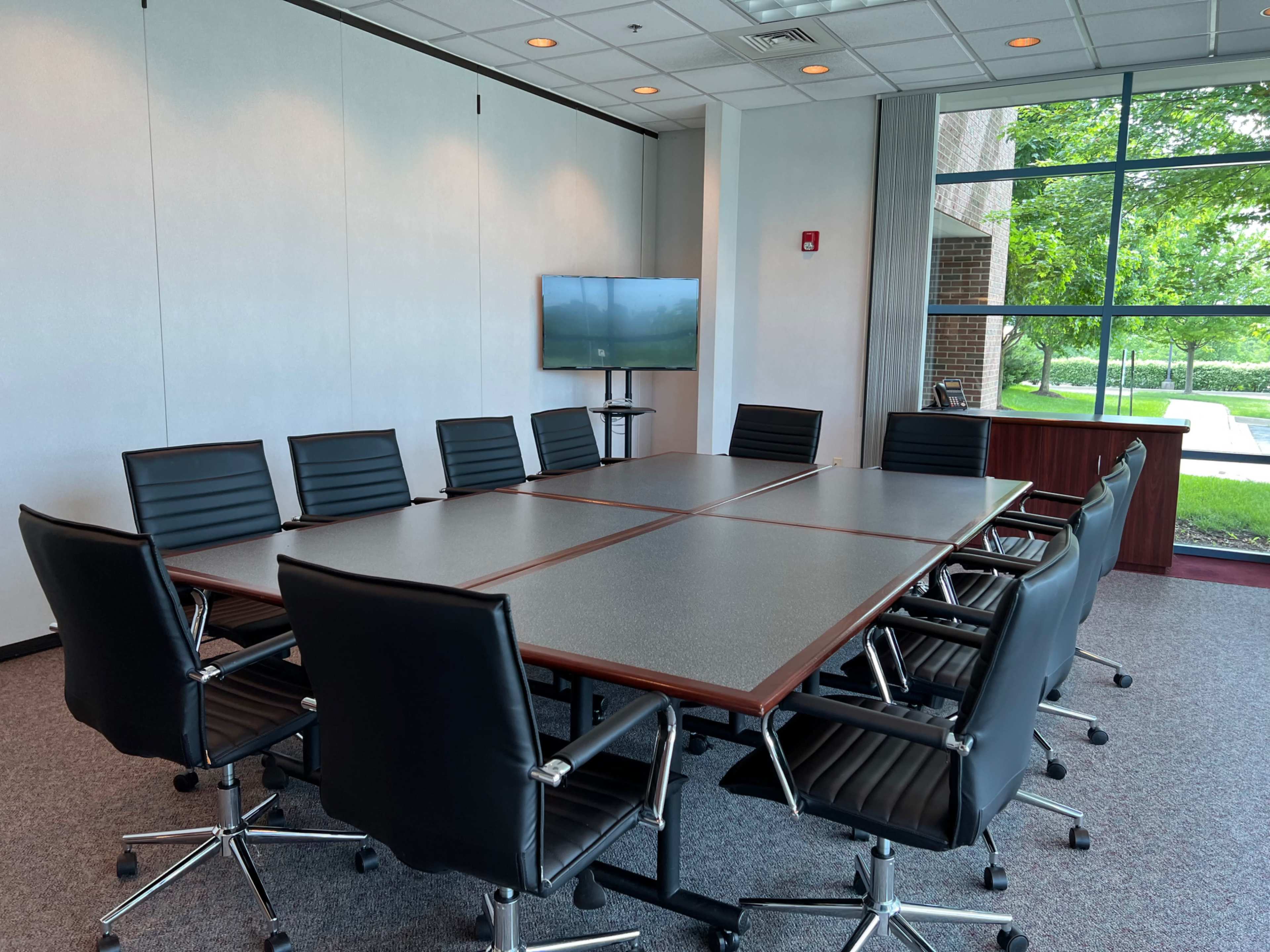 A conference room features a large rectangular table surrounded by eight black leather chairs and a screen mounted on the wall.