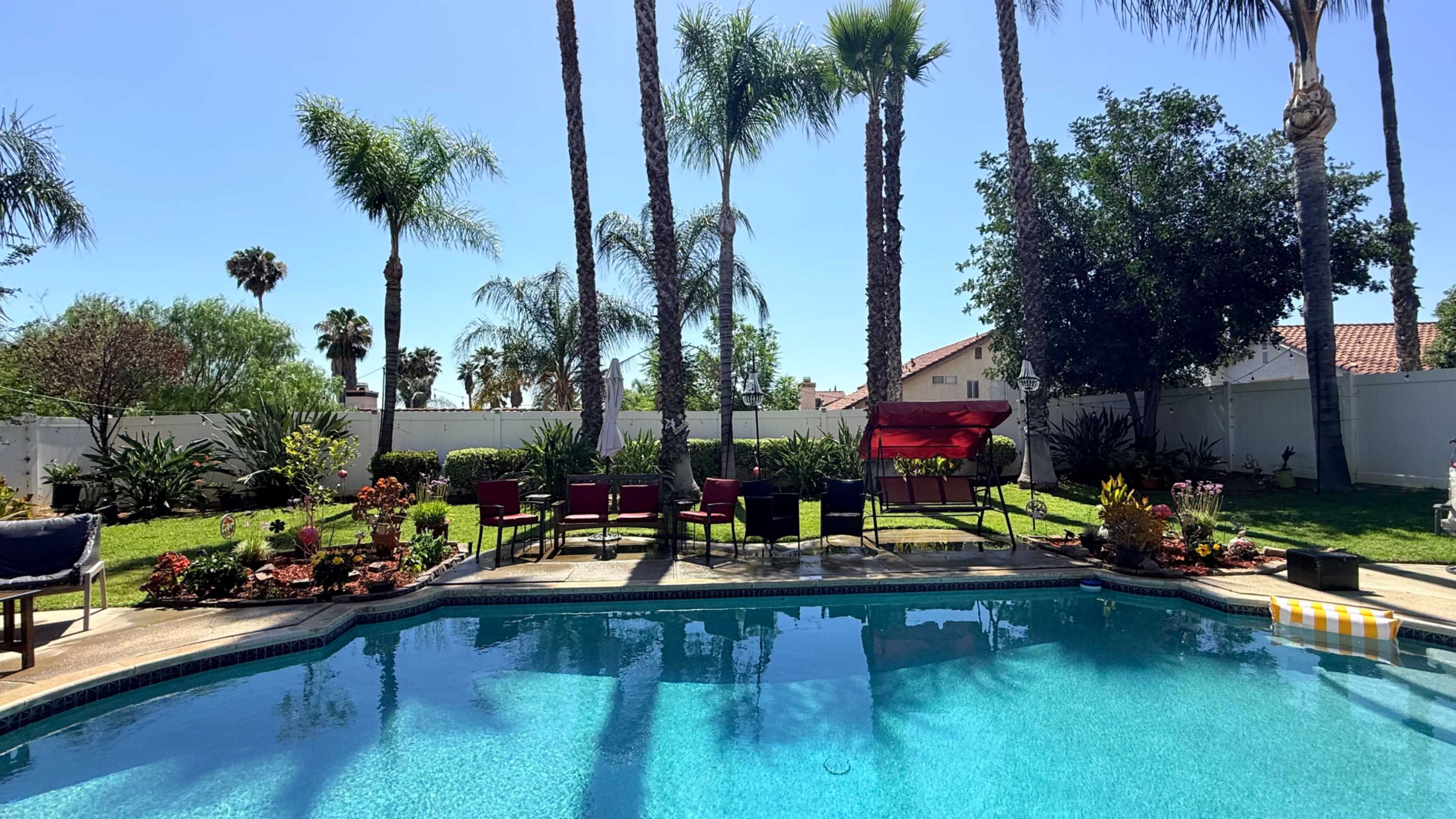 The image shows a brightly lit swimming pool surrounded by palm trees, lounge chairs, and a garden with colorful plants.