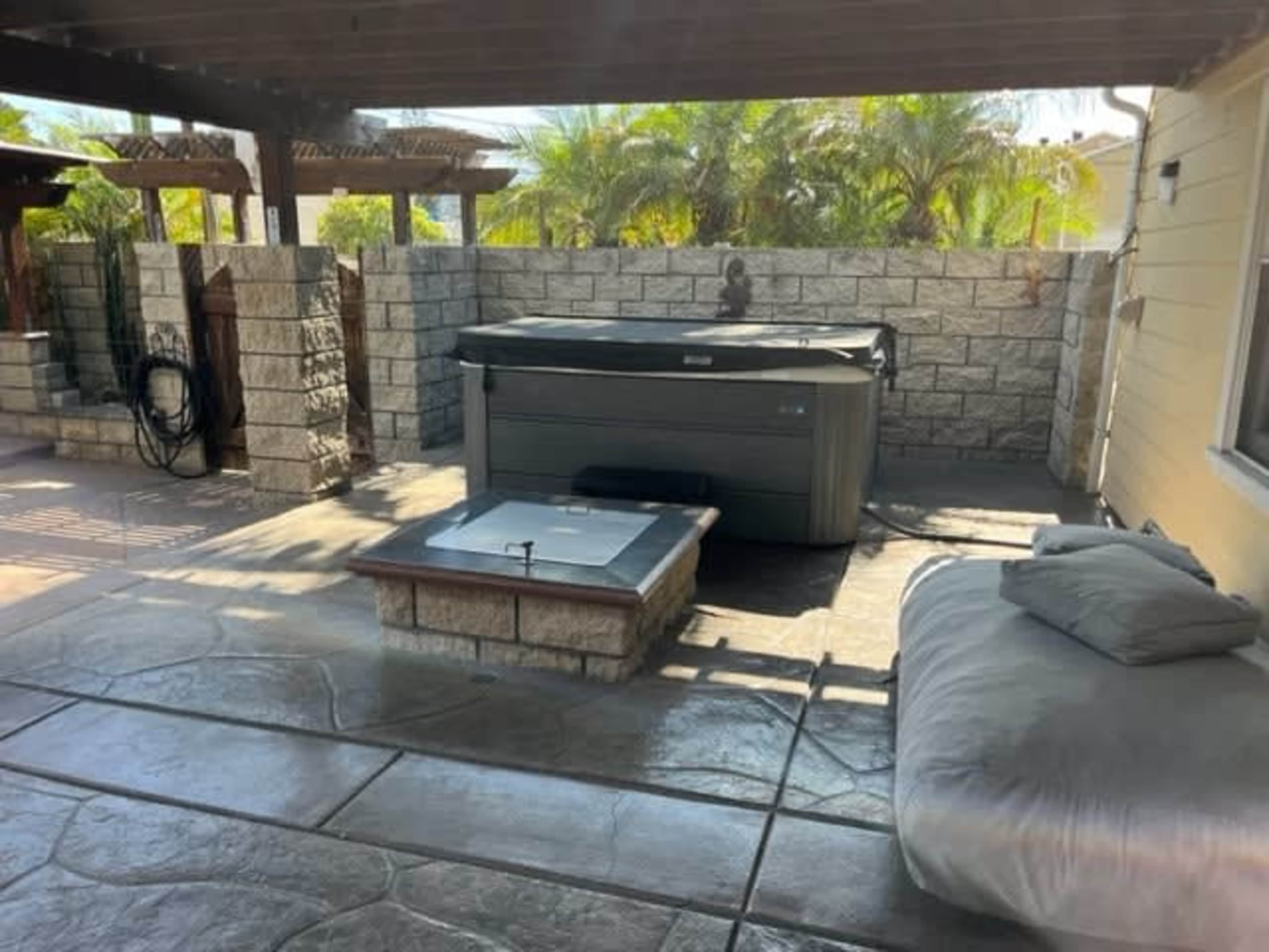 The image shows a patio area with a hot tub, a fire pit, and a large cushion beside a wall of stone and greenery in the background.