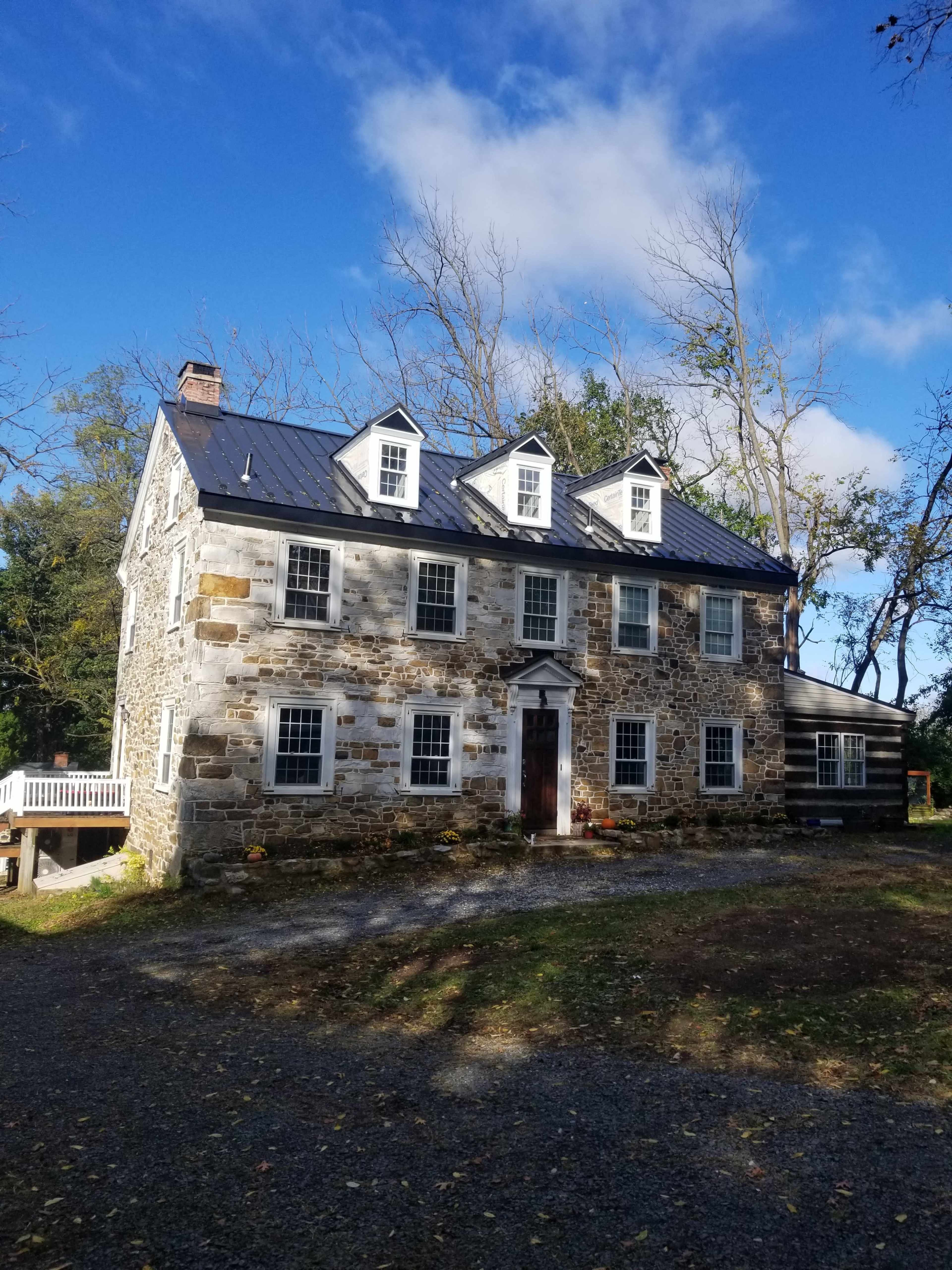 A stone house with a slate roof and multiple windows stands amidst a gravel driveway and trees under a partly cloudy sky.