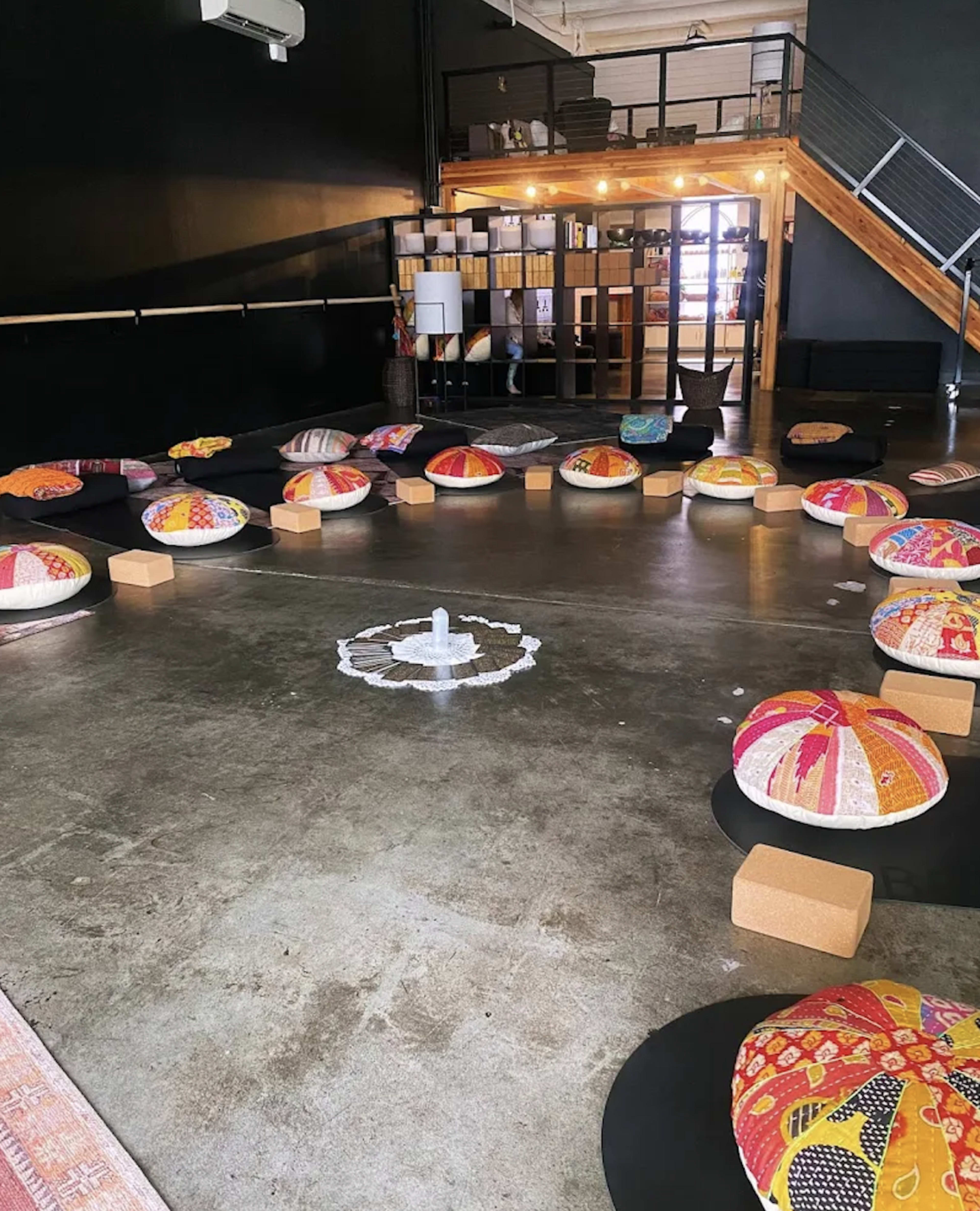 The image shows a spacious room with a circular arrangement of colorful padded stools surrounding a central design on the floor, with wooden stairs visible in the background.