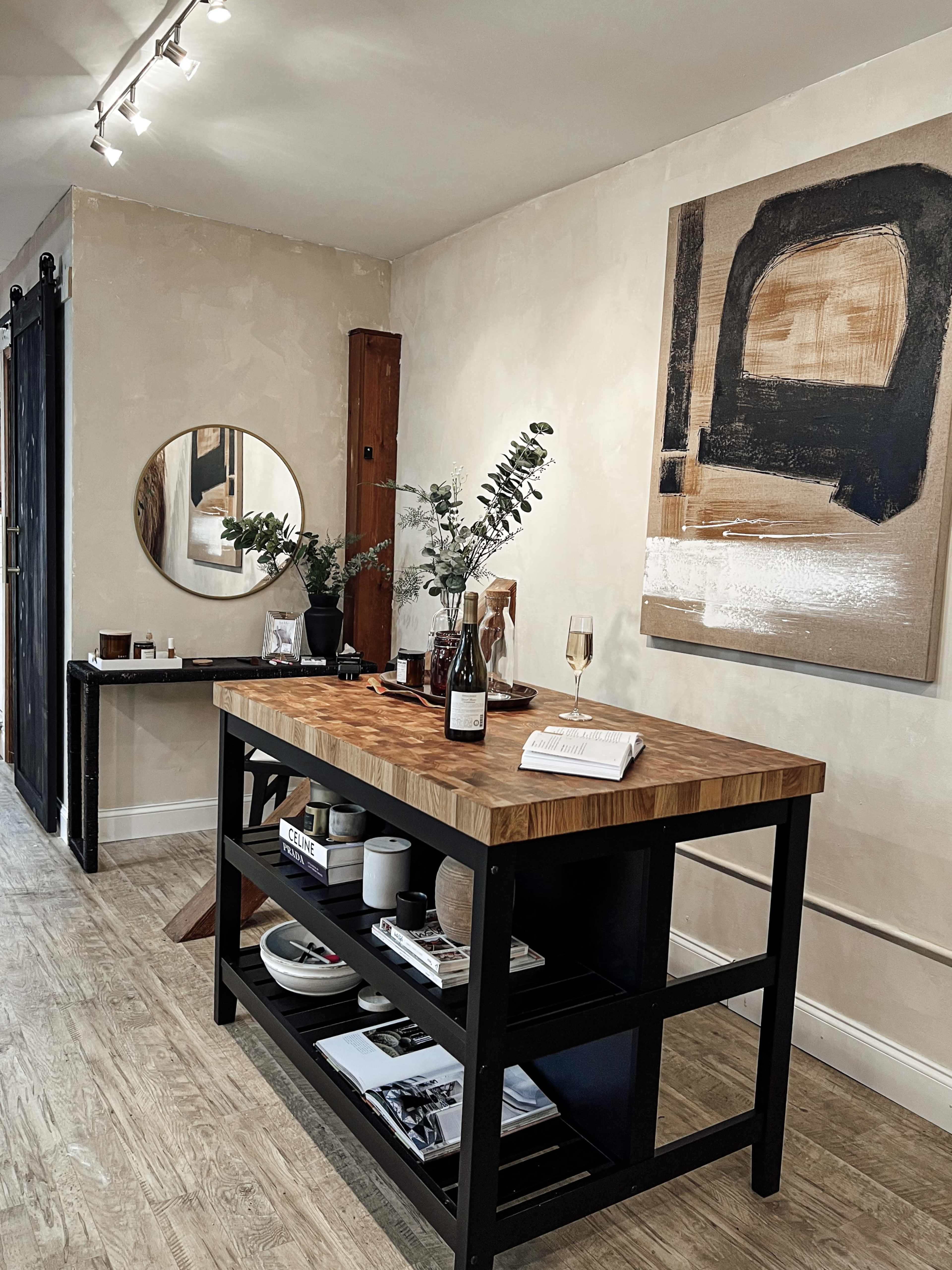 A wooden island table sits in a modern room adorned with decorative artwork, a round mirror, and various decorative items on a shelf below.