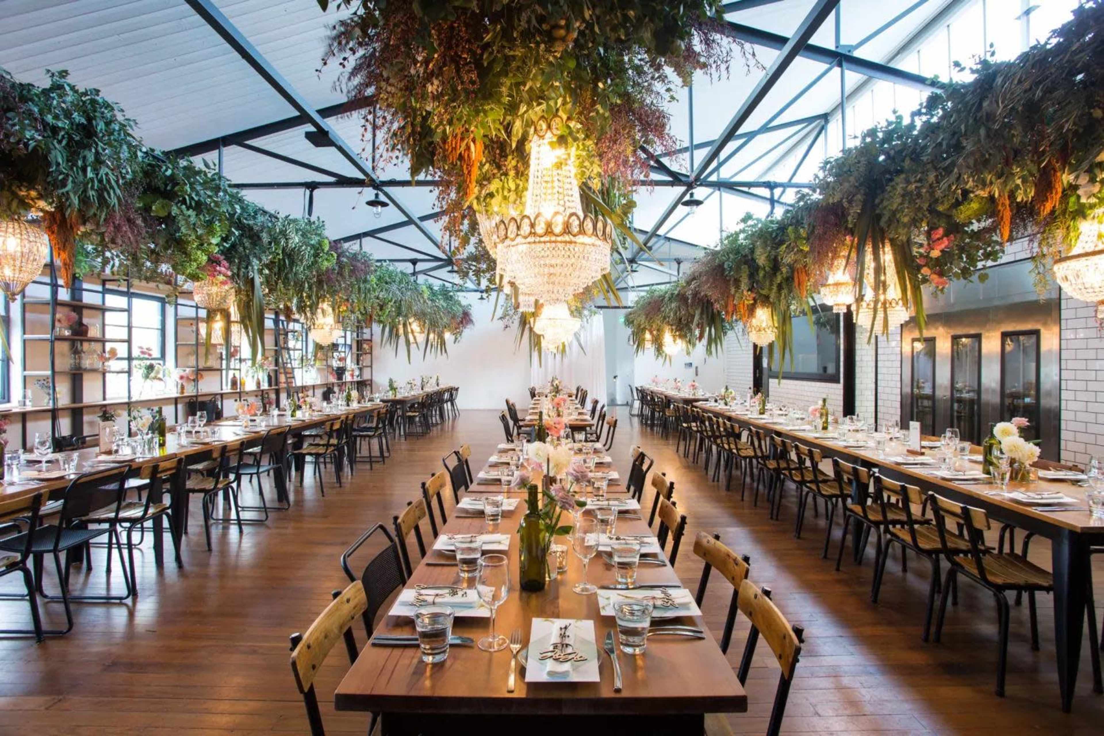 A large dining area with long wooden tables set with plates and glasses, surrounded by hanging greenery and chandeliers.