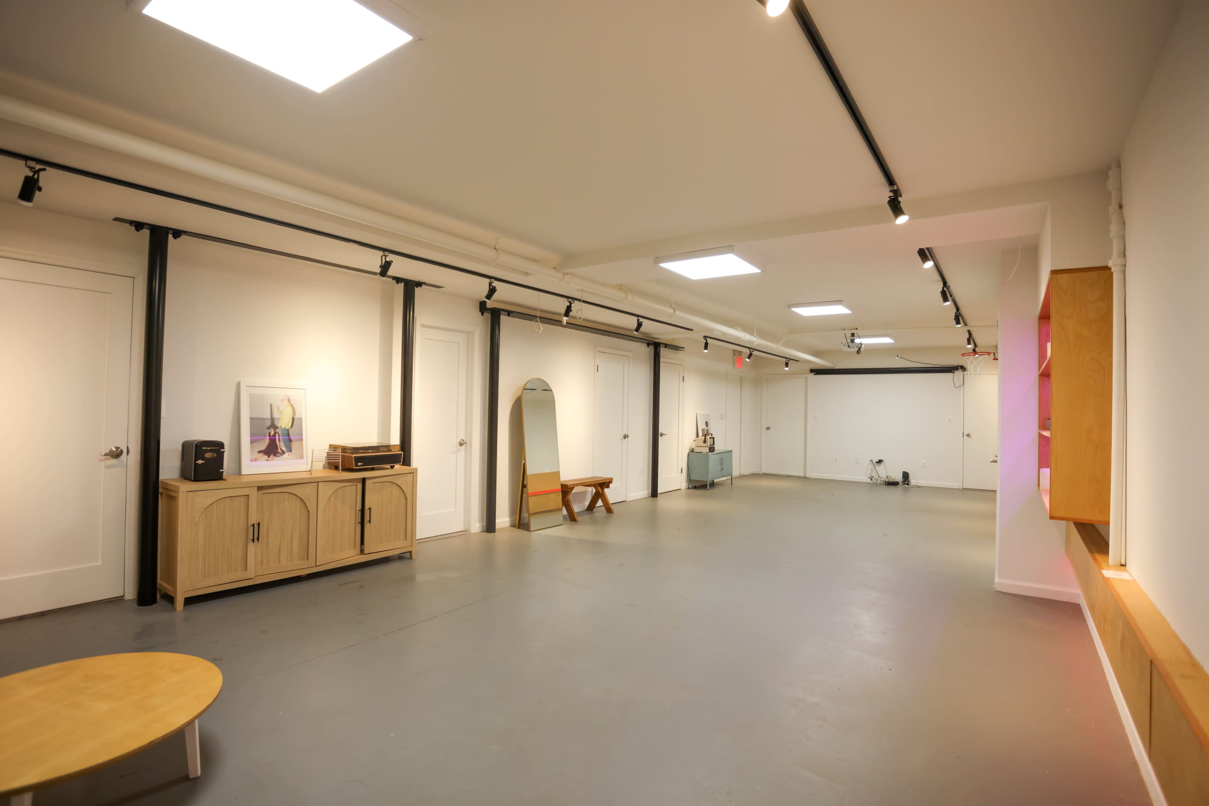 The image shows a spacious, well-lit room with gray flooring, white walls, and minimalist decor including a cabinet, a mirror, and a small table.