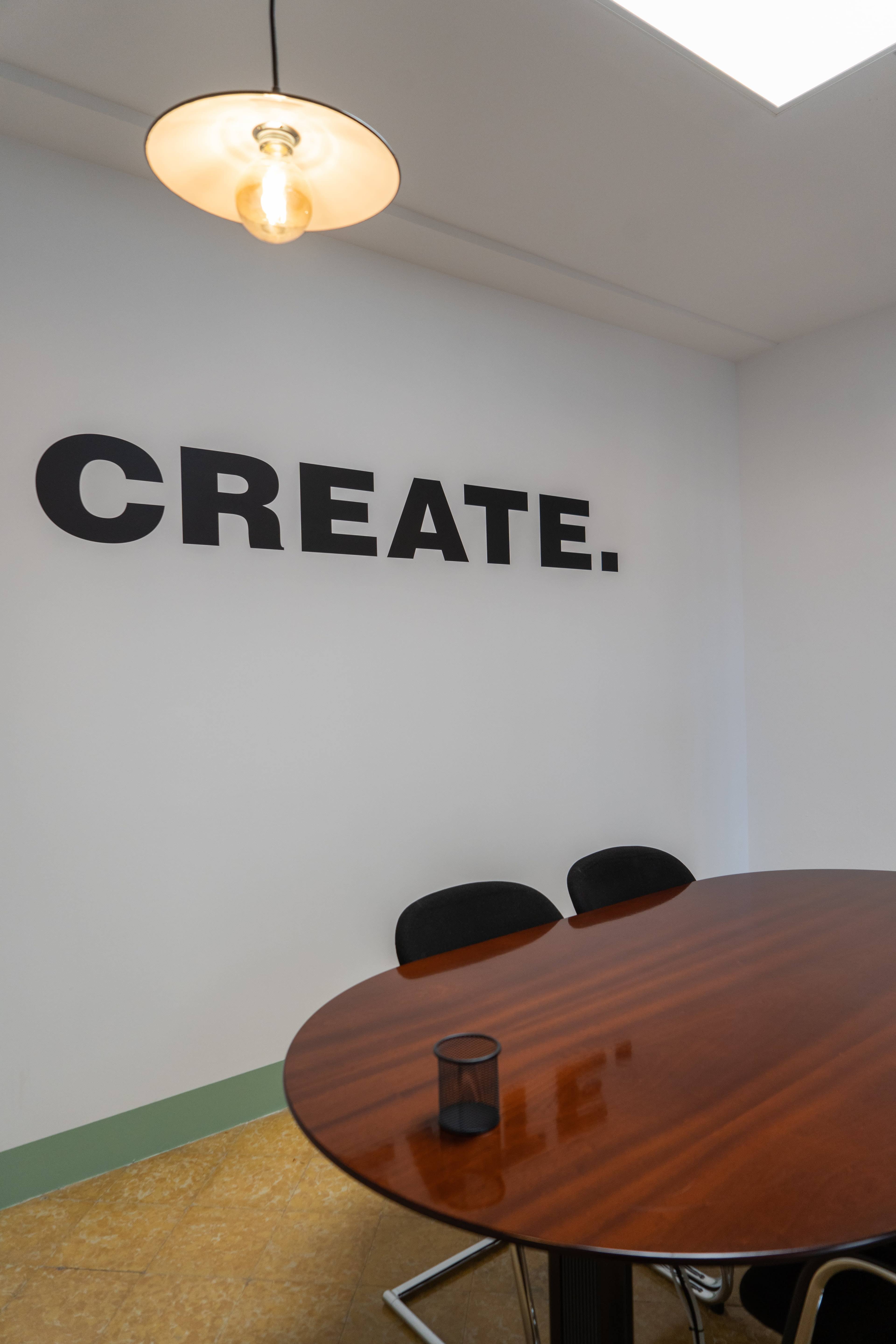 A round wooden table with black chairs is situated in a room featuring a large wall sign that reads "CREATE."