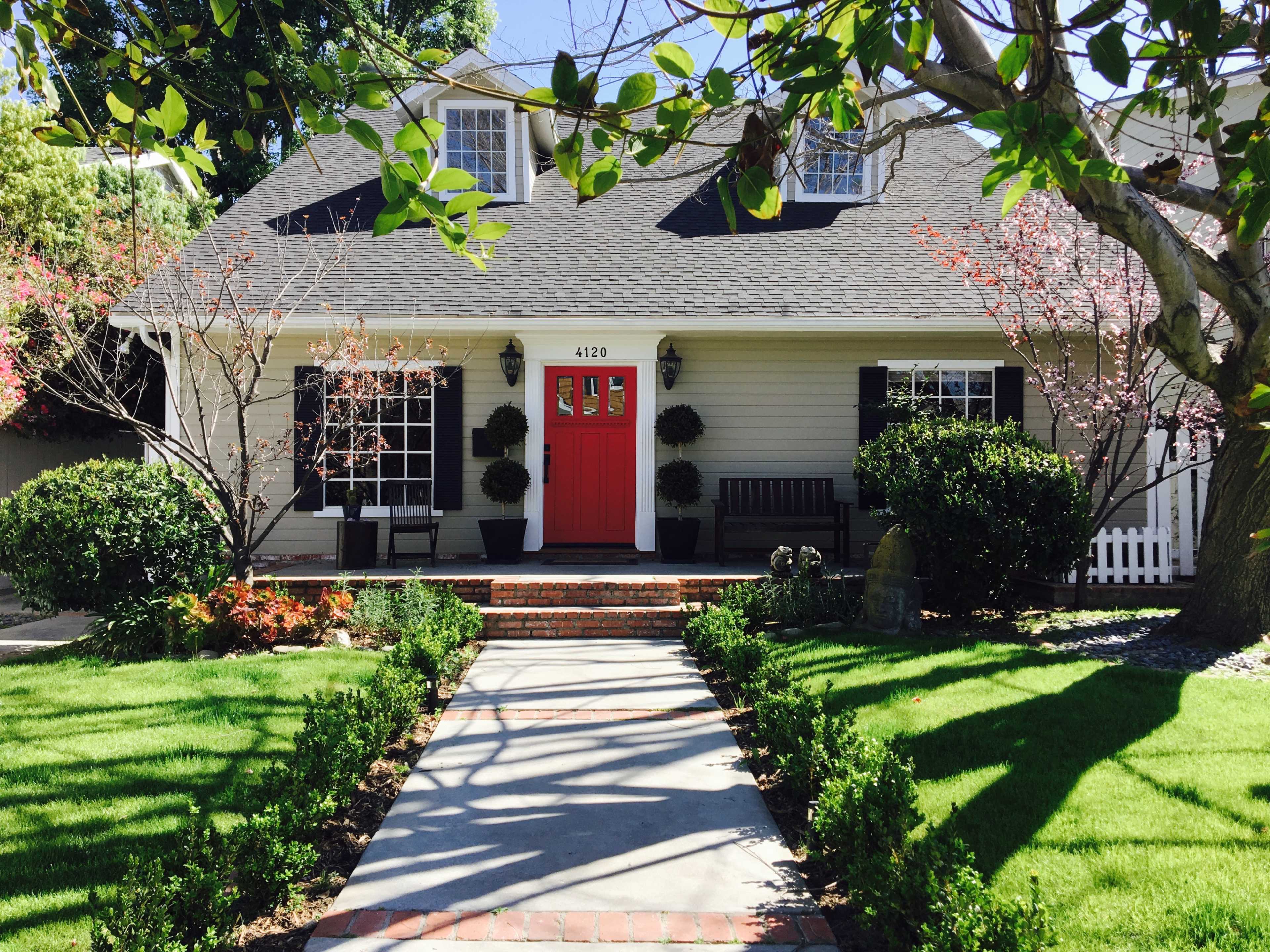 A single-story house with a gray exterior, a red front door, and neatly trimmed greenery lines a brick walkway leading to the entrance.