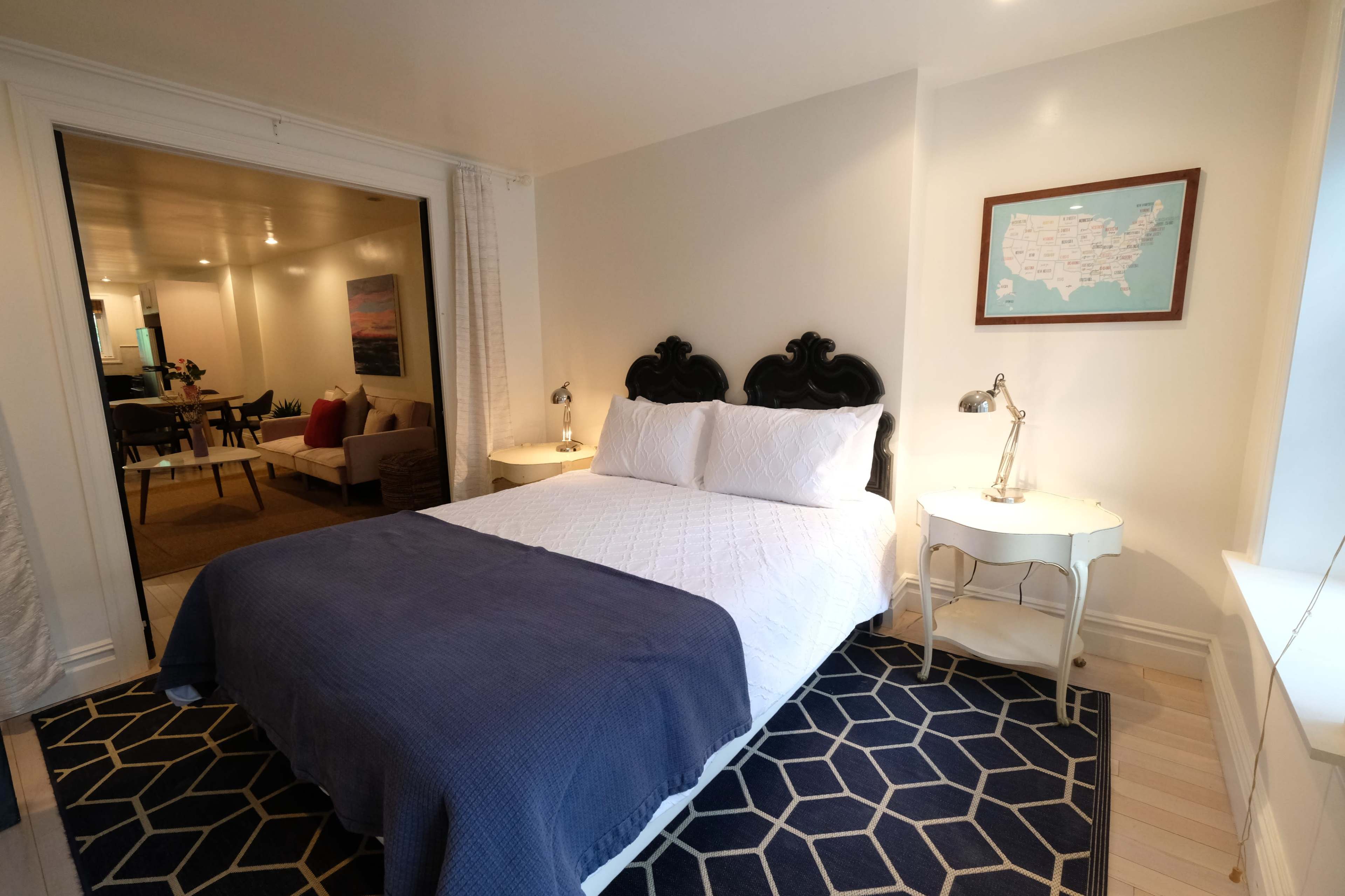 The image shows a well-lit bedroom featuring a black upholstered headboard, a white bed with blue bedding, and a nightstand with lamps, alongside an open doorway leading to a living area.