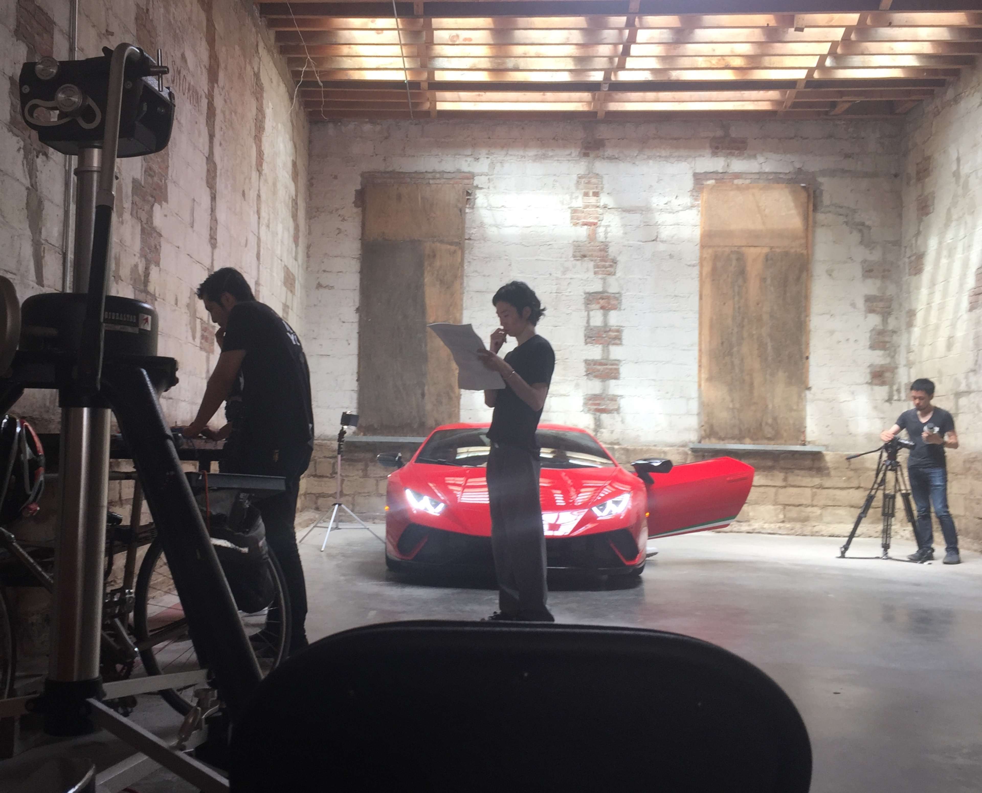 Three individuals are engaged in different activities in a well-lit garage, with a red sports car prominently featured in the background.
