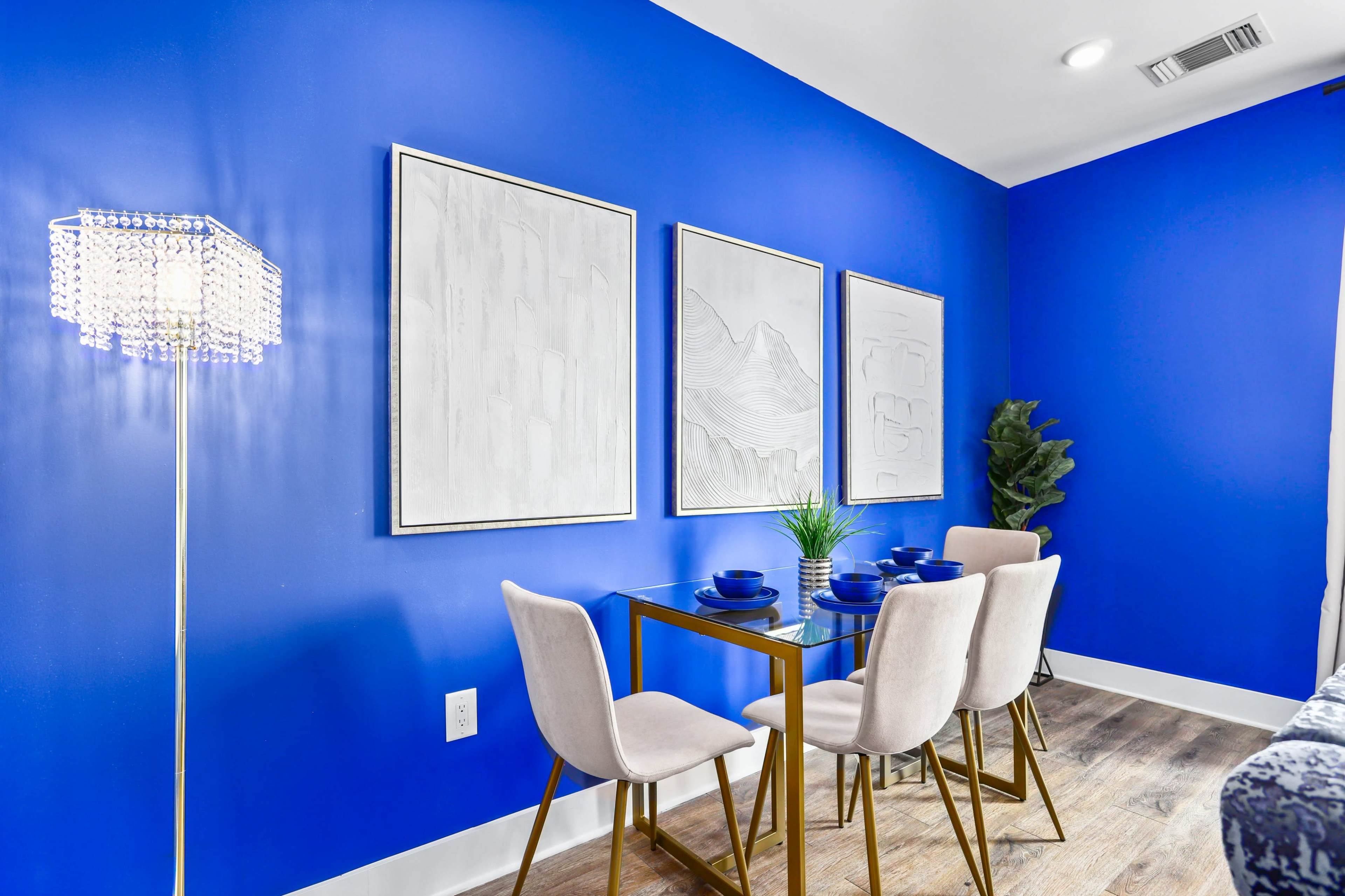 A modern dining area features a blue accent wall, a glass table with four chairs, and three framed artworks above the table.