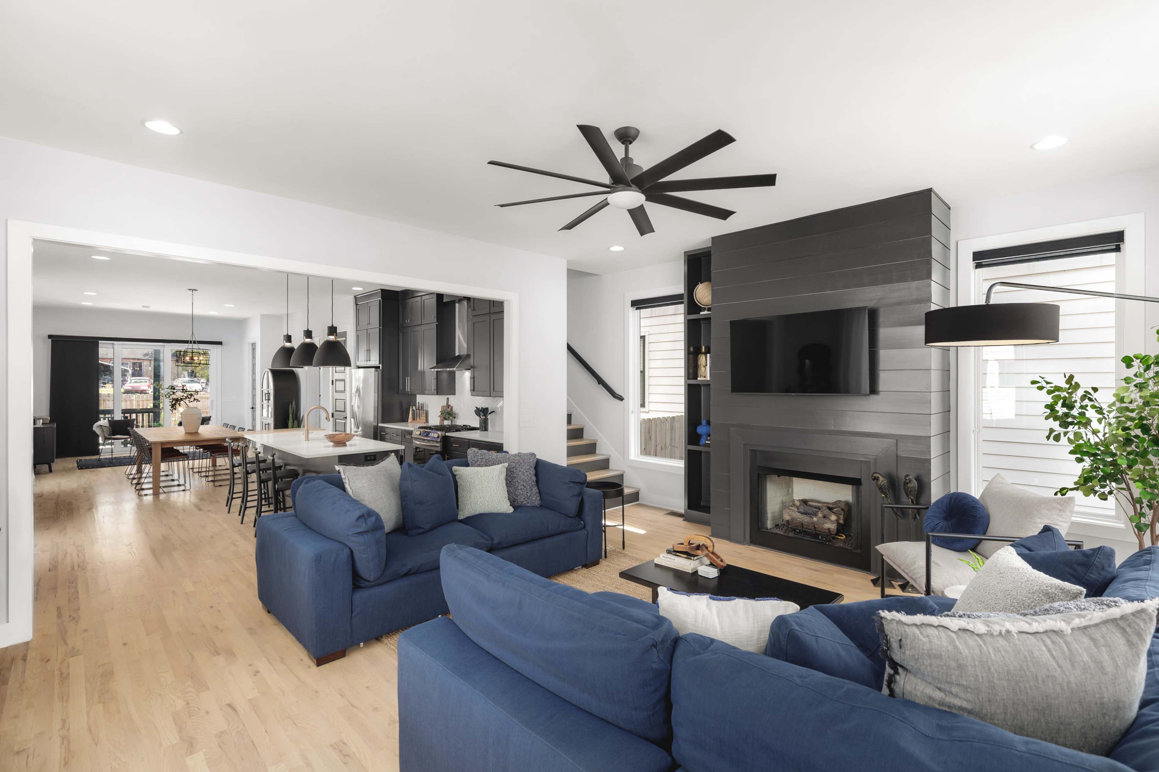 A modern living room with a blue sectional sofa, a fireplace, and a large ceiling fan, connected to a dining area in the background.
