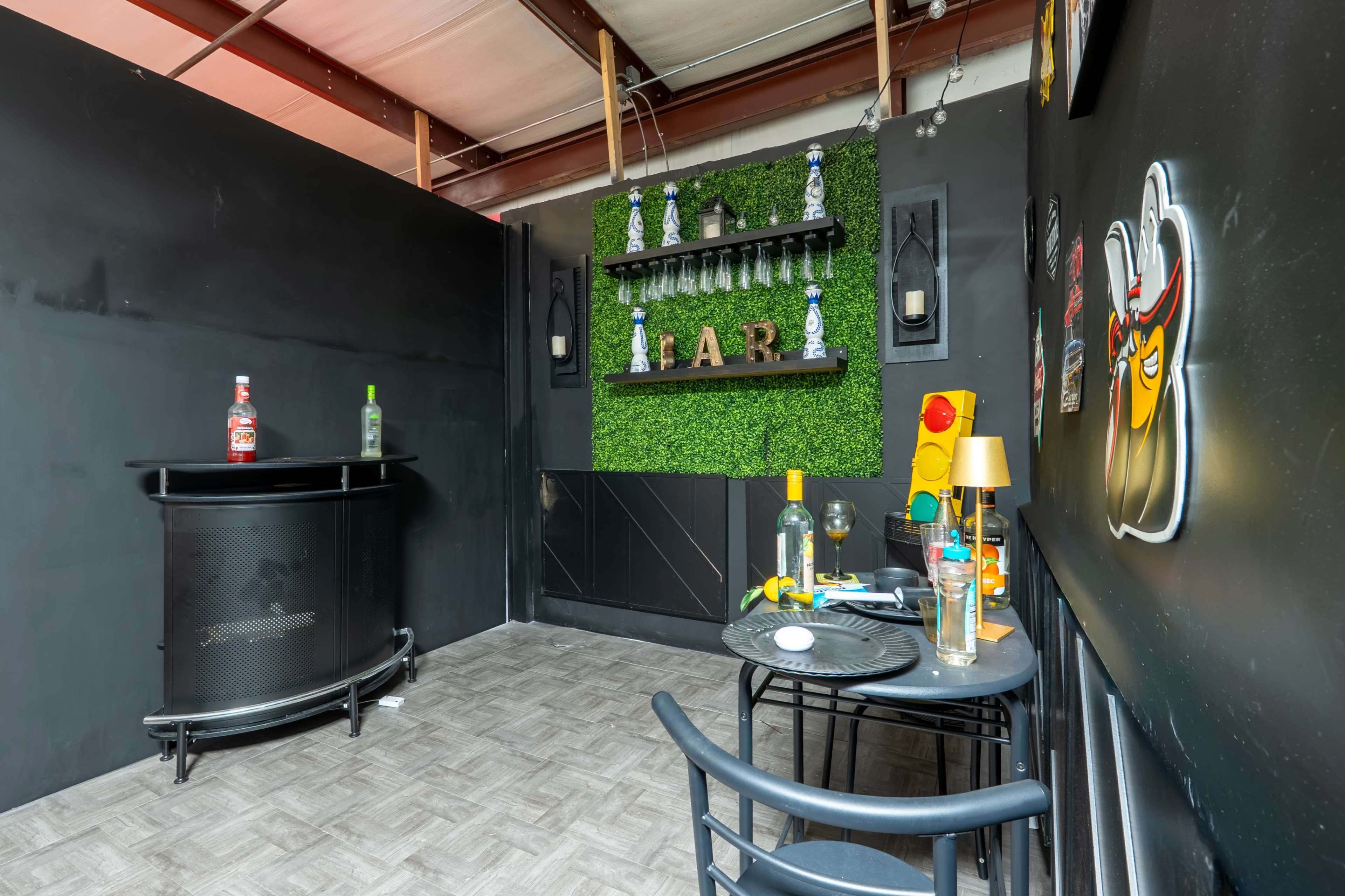 The image shows a small bar area with a dark wall, a black circular bar cart, two chairs, and decorative shelves featuring bottles and greenery.