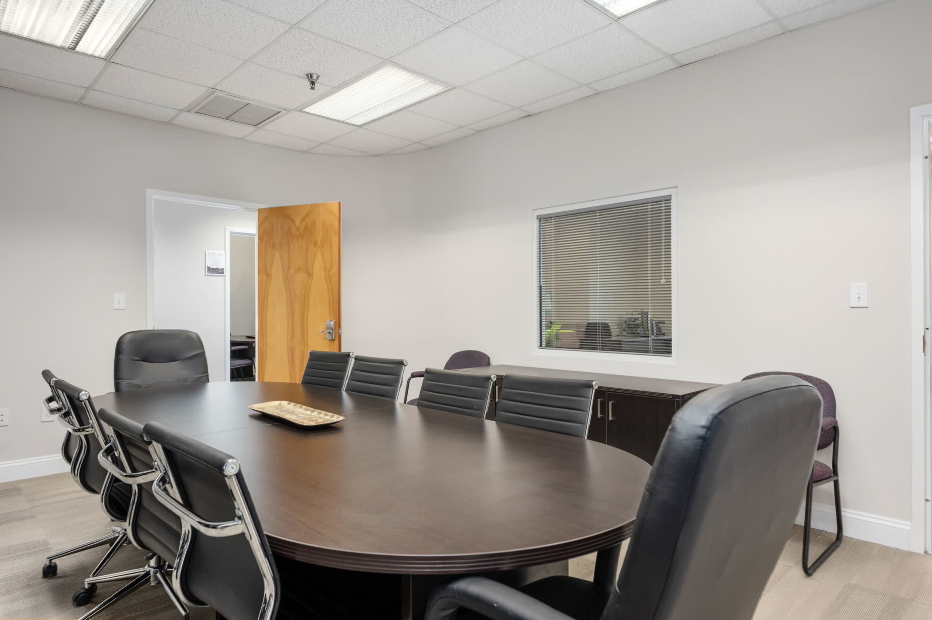 A conference room features a large oval table surrounded by black chairs, with a window and a door leading to another room.