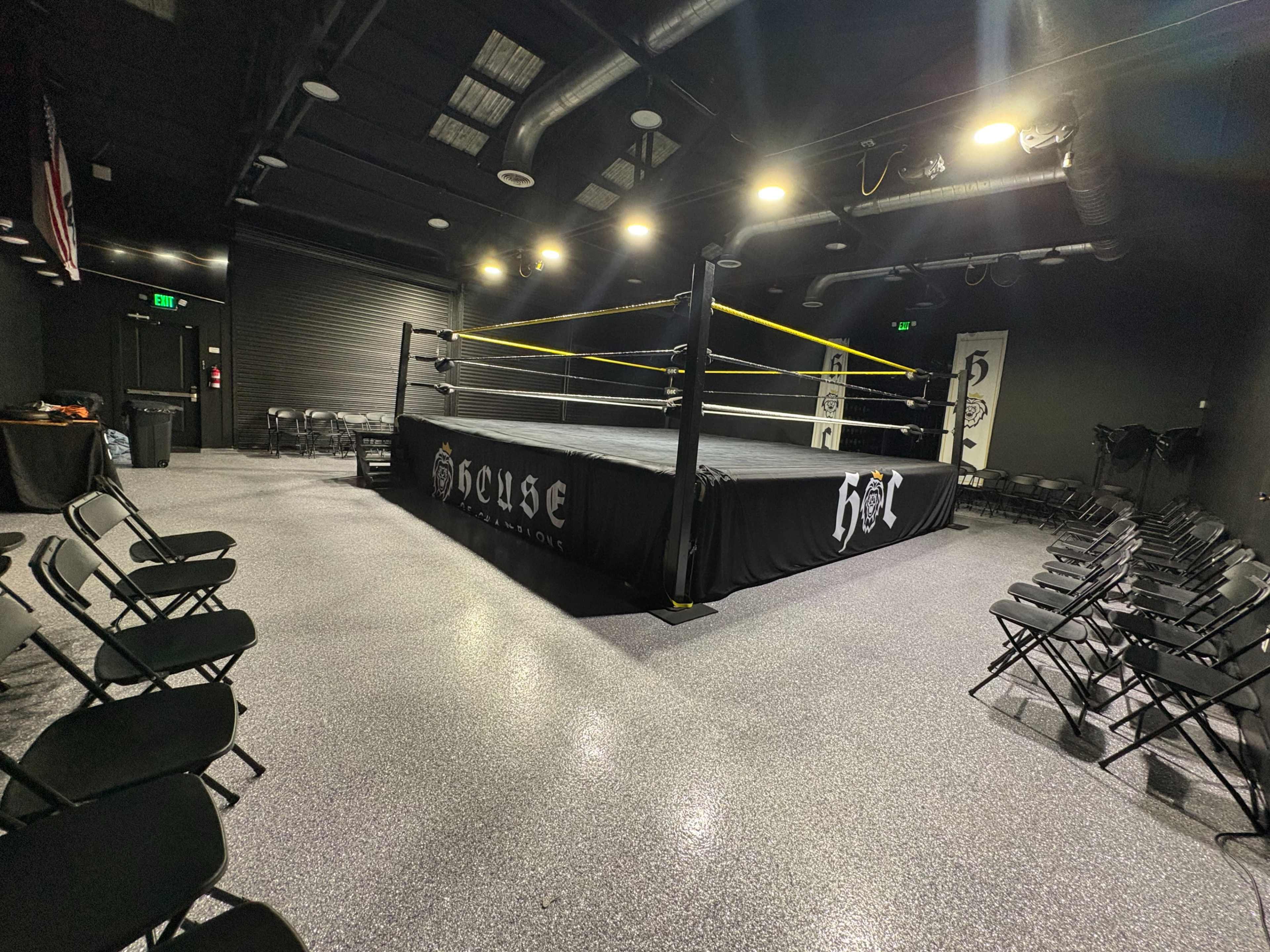 The image shows a professional wrestling ring in a dimly lit training facility, surrounded by black folding chairs and featuring a black and yellow aesthetic.