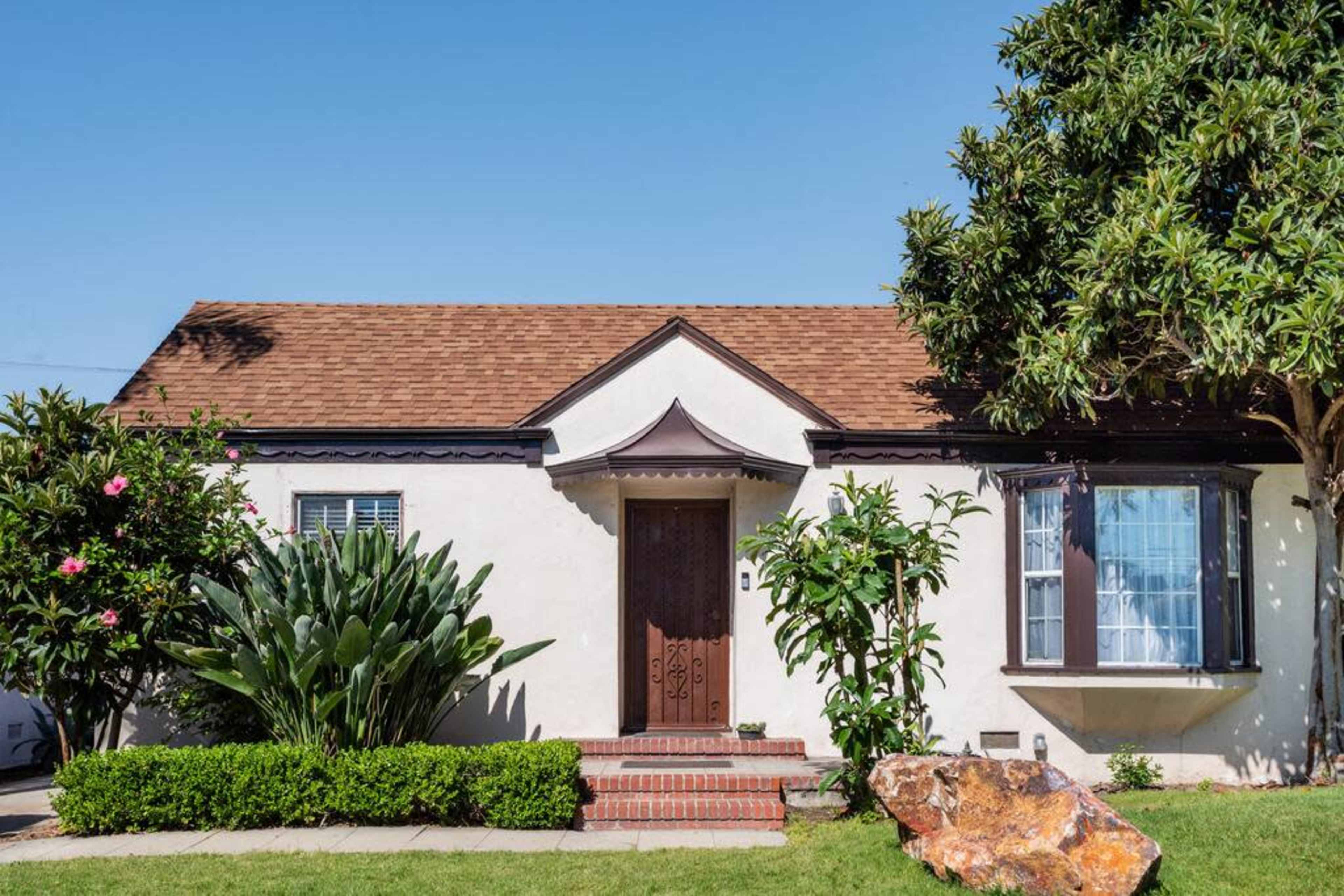 A single-story house with a brown shingle roof, a front porch, and landscaped greenery including shrubs and plants.
