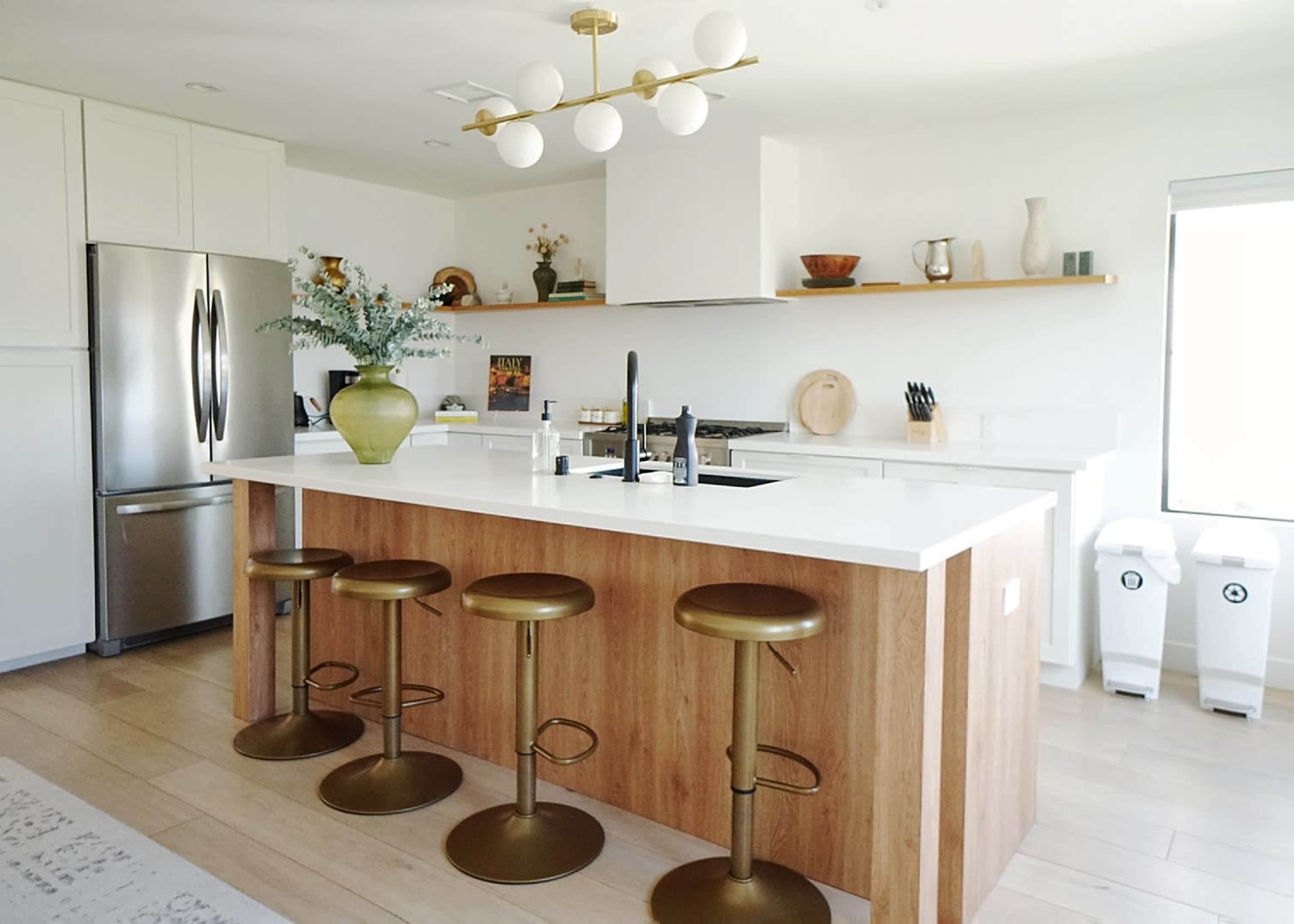 The image shows a modern kitchen featuring a central island with four gold bar stools, a stainless steel refrigerator, and a minimalist decor of vases and kitchenware on open shelves.