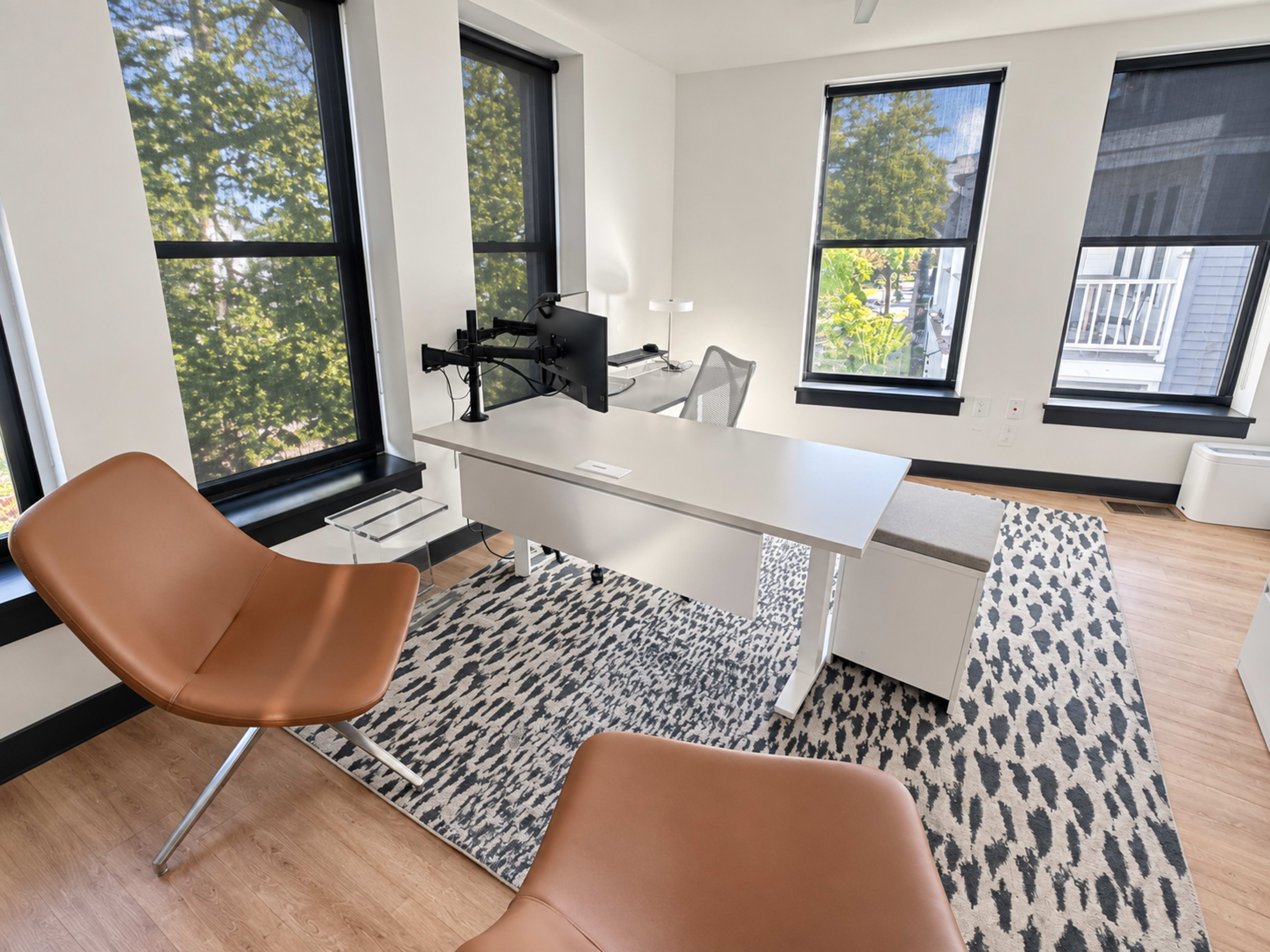 The image shows a modern office space featuring a desk with a computer monitor, a comfortable chair, and large windows allowing natural light.