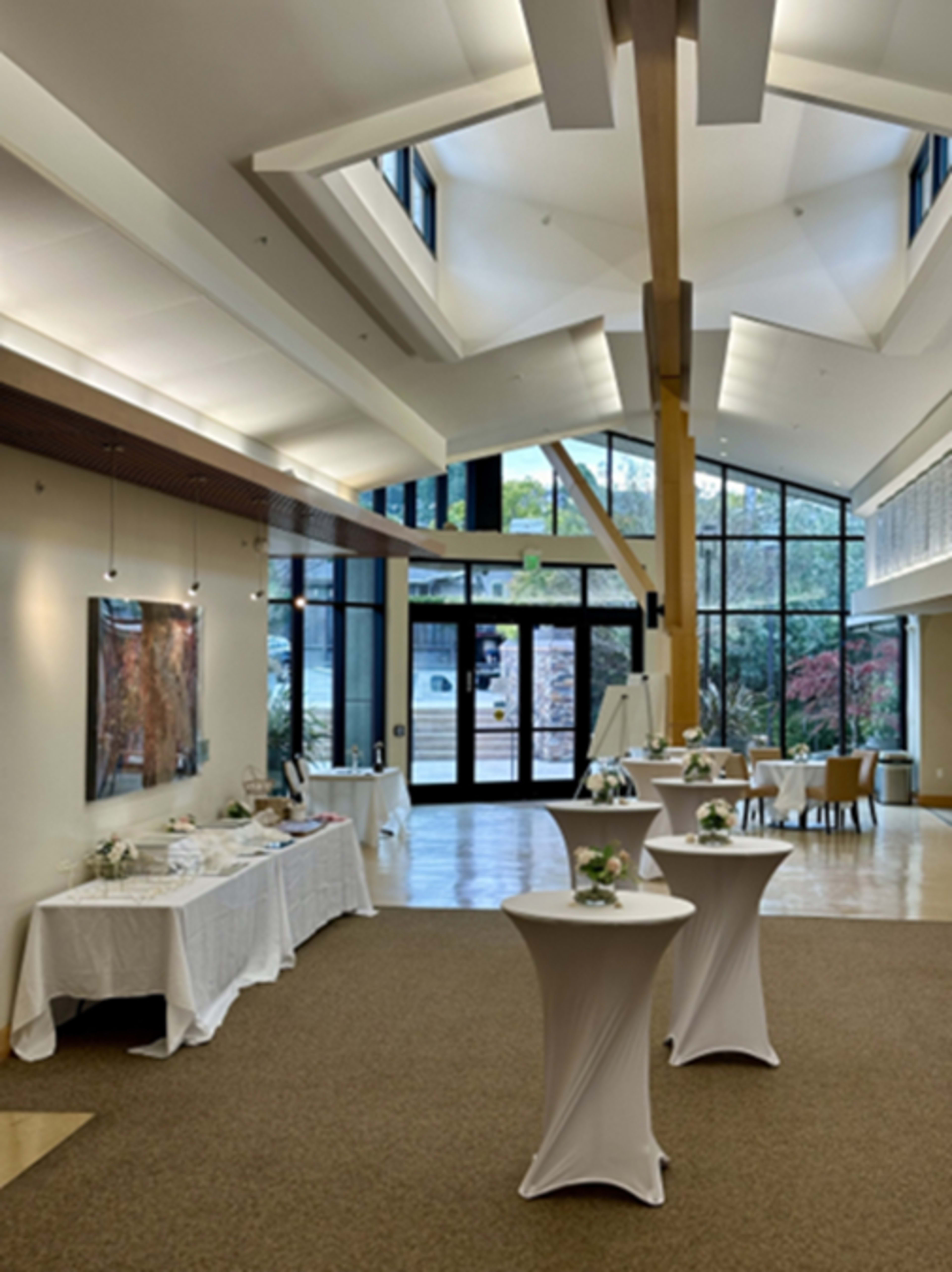 The image shows a spacious, well-lit interior of a venue with several round tables and a large glass entrance, featuring white table linens and floral centerpieces.